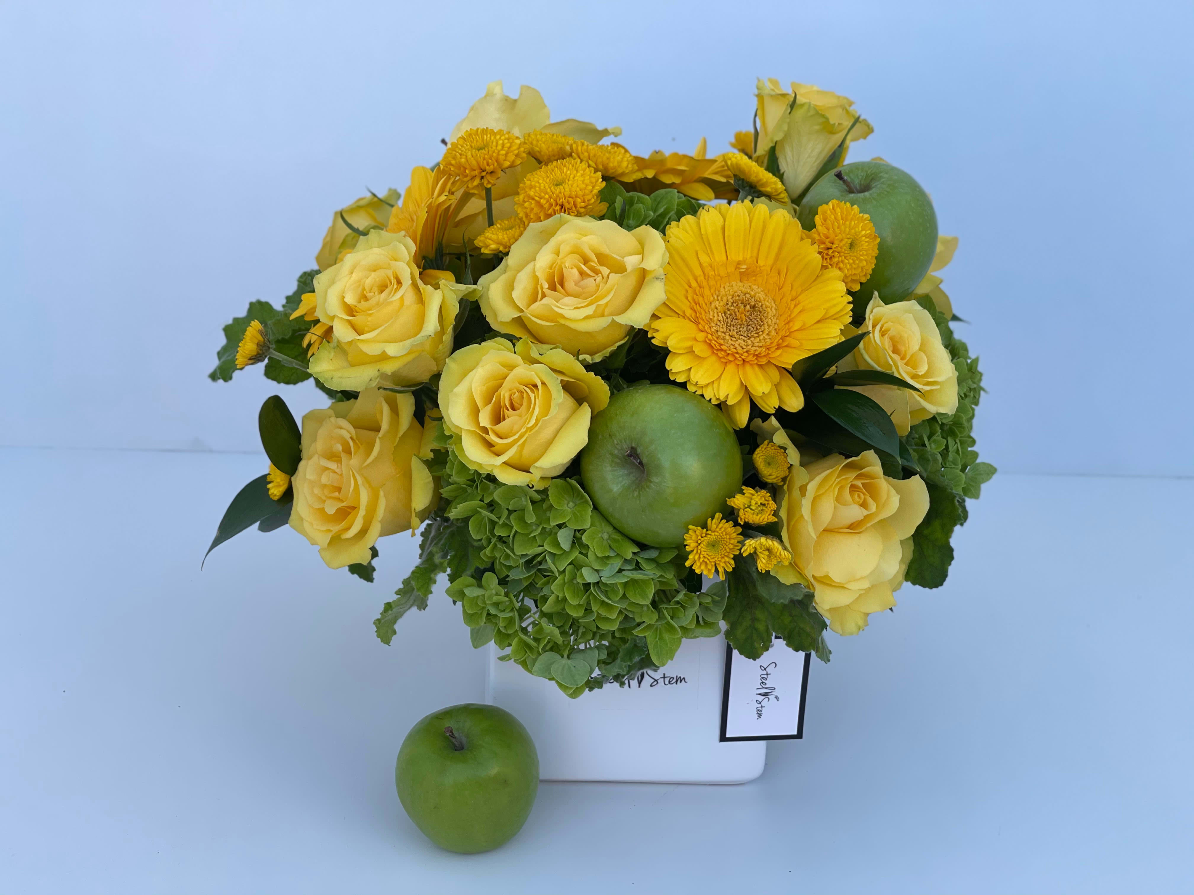 Green Apple - A green and yellow chic arrangement of yellow roses, emerald green hydrangeas, yellow gerbera daisies, yellow chrysanthemum, a hint of green leafs and complimented with green apples that is hand-designed inside of white square ceramic vase.