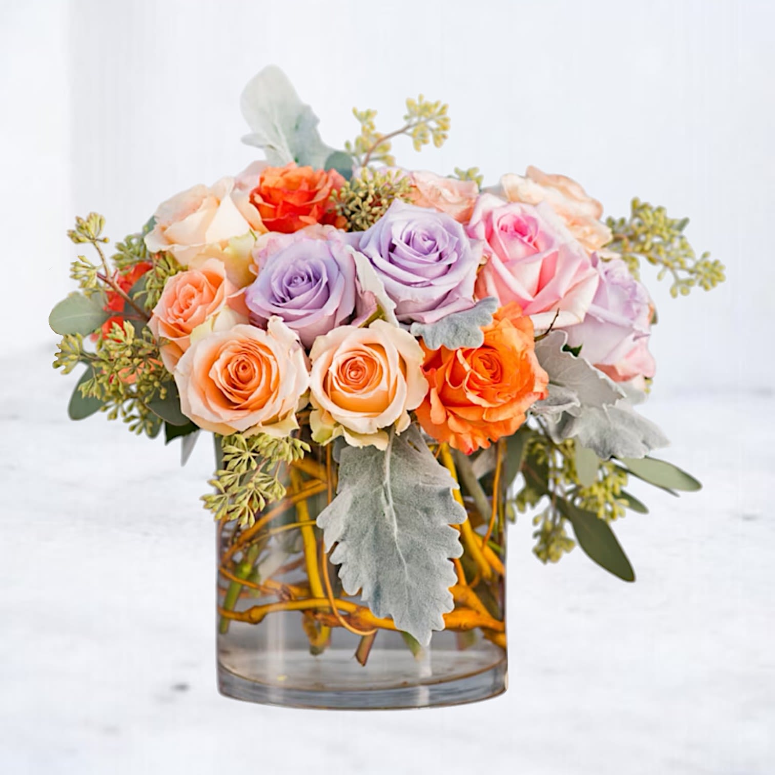 Pastel Sorbet - Elevate any moment with Pastel Sorbet, a stunning bouquet blending soft pastels with a fiery touch. Featuring lush lavender, peach, and apricot roses, accented by delicate dusty miller and wisps of textural greenery, this arrangement is both ethereal and bold. The clear vase showcases natural vine accents, adding an organic elegance. Perfect for birthdays, celebrations, or as an artistic floral gift, this design brings warmth and sophistication in one breathtaking display.