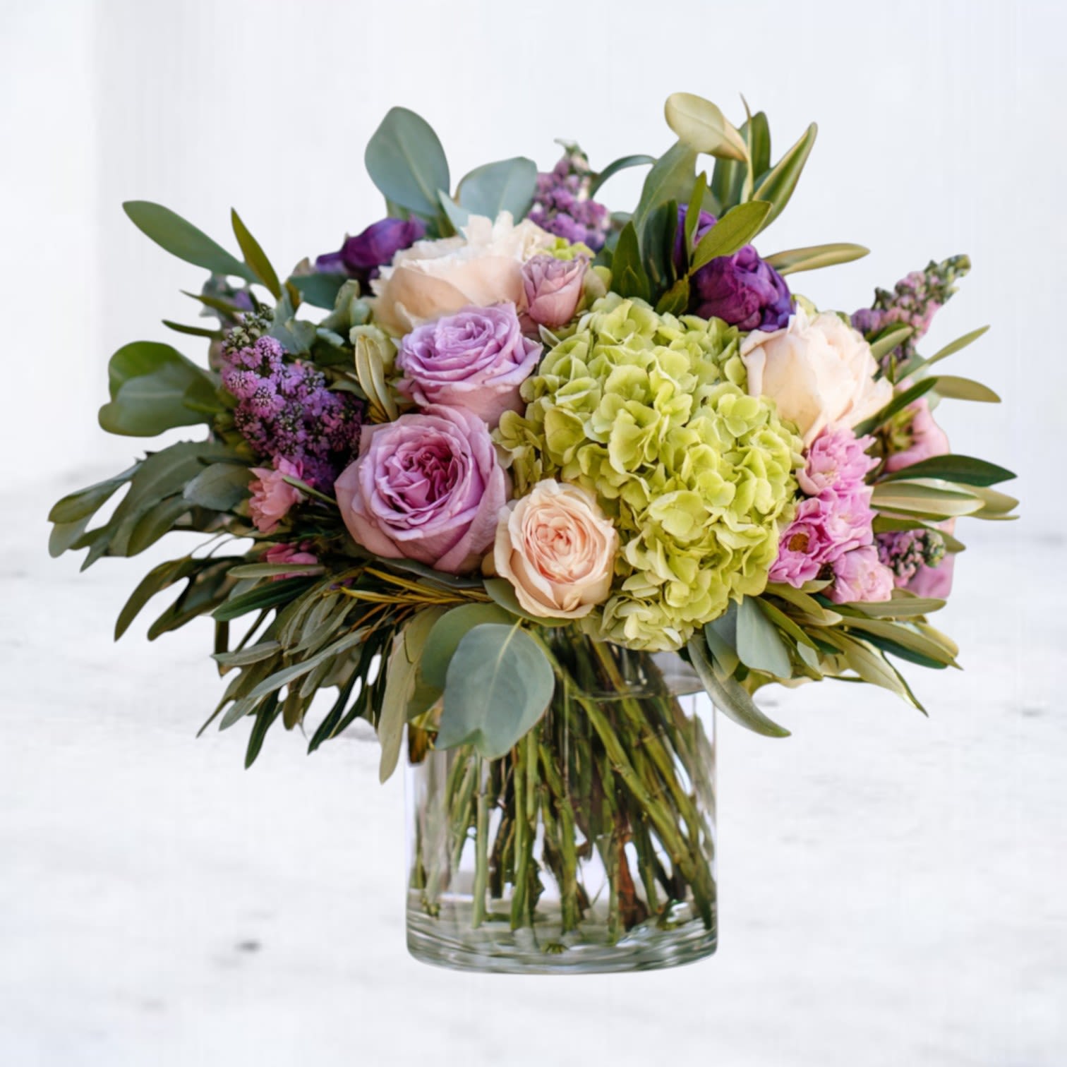 Lavender Composition - Lavender Composition blends soft lavender roses with green hydrangea, blush carnations, and fresh seasonal greenery in a clear glass cylinder vase. The layered blooms create natural movement while the muted palette keeps the design balanced and effortless.  Light yet dimensional, this arrangement feels thoughtful without being formal — perfect for sending care, marking a quiet milestone, or brightening someone’s space with understated color.  Available for same-day flower delivery in Houston and surrounding areas.