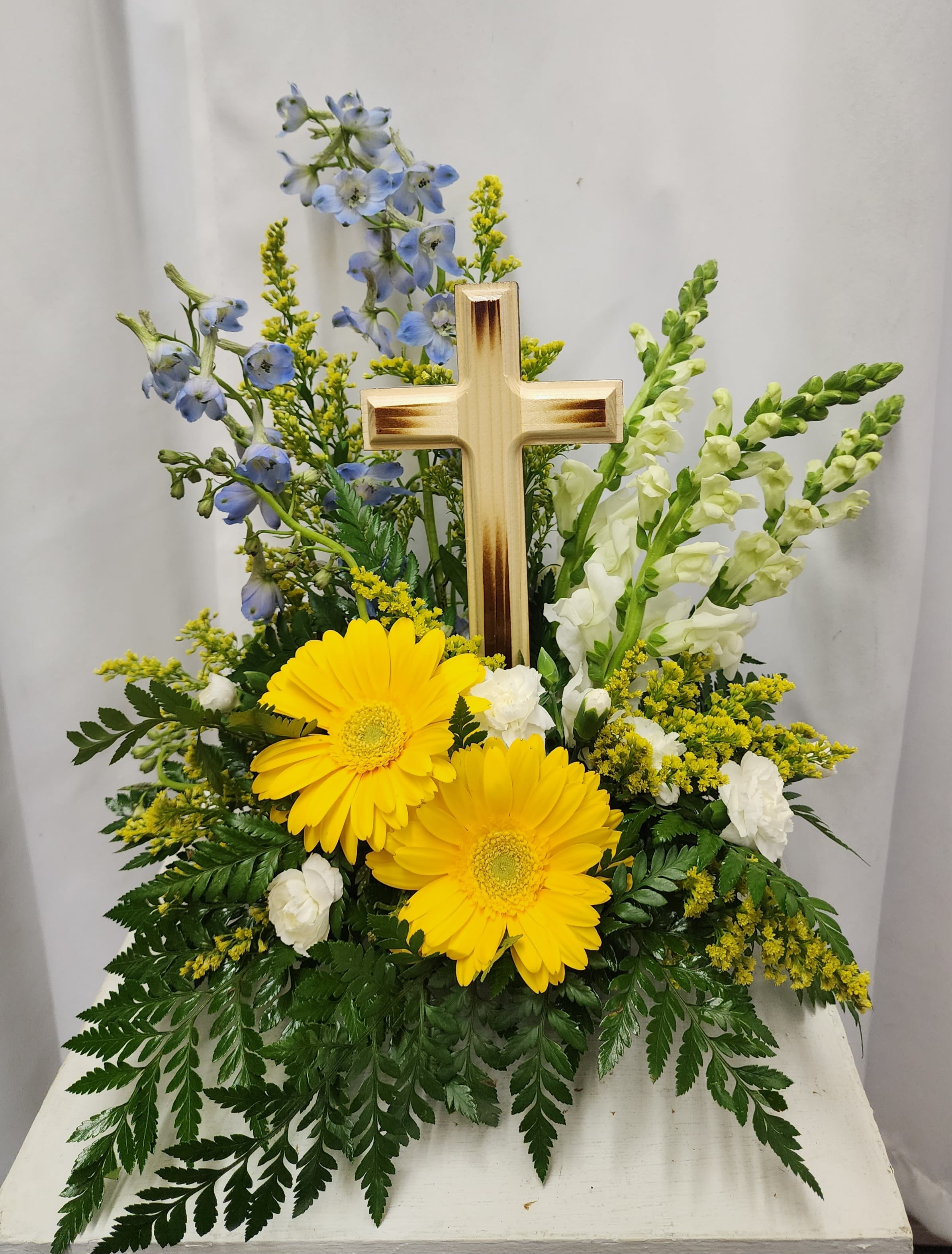 Sunshine on the Horizon - Medium Cross - Bright and colorful yet soft and peaceful describes this elegant sympathy arrangement.  The handcrafted 7" wooden cross keepsake is assembled with exceptional care and attention to detail. The flowers include yellow gerbera daisies, light blue delphinium, white snapdragons, mini carnations and yellow solidago. Can be made with various color gerbs if desired. 
