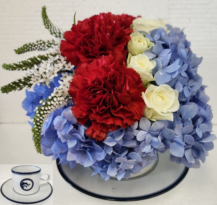 The Spirit of America - In a keepsake Coffee Cup with Saucer this patriotic arrangement has Red Carnations, White Spray Roses, White Gooseneck Cone Flowers arranged in a bed of Blue Hydrangeas.