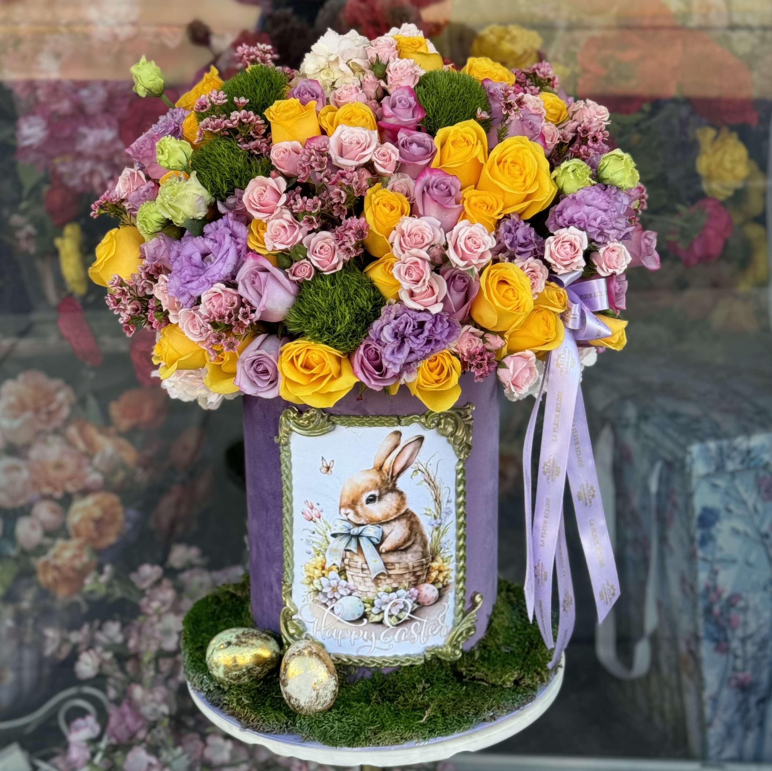 Easter Bunny’s Garden - A delightful Easter arrangement bursting with the colors of spring! This stunning bouquet features a rich blend of yellow, pink, and lavender roses, complemented by lush greenery and delicate florals. The elegant purple hatbox, adorned with a charming vintage-style Easter bunny illustration, adds a whimsical and festive touch. Finishing details like golden Easter eggs and decorative ribbons make this design truly special.  Perfect for gifting, decorating an Easter table, or bringing seasonal cheer into any space!