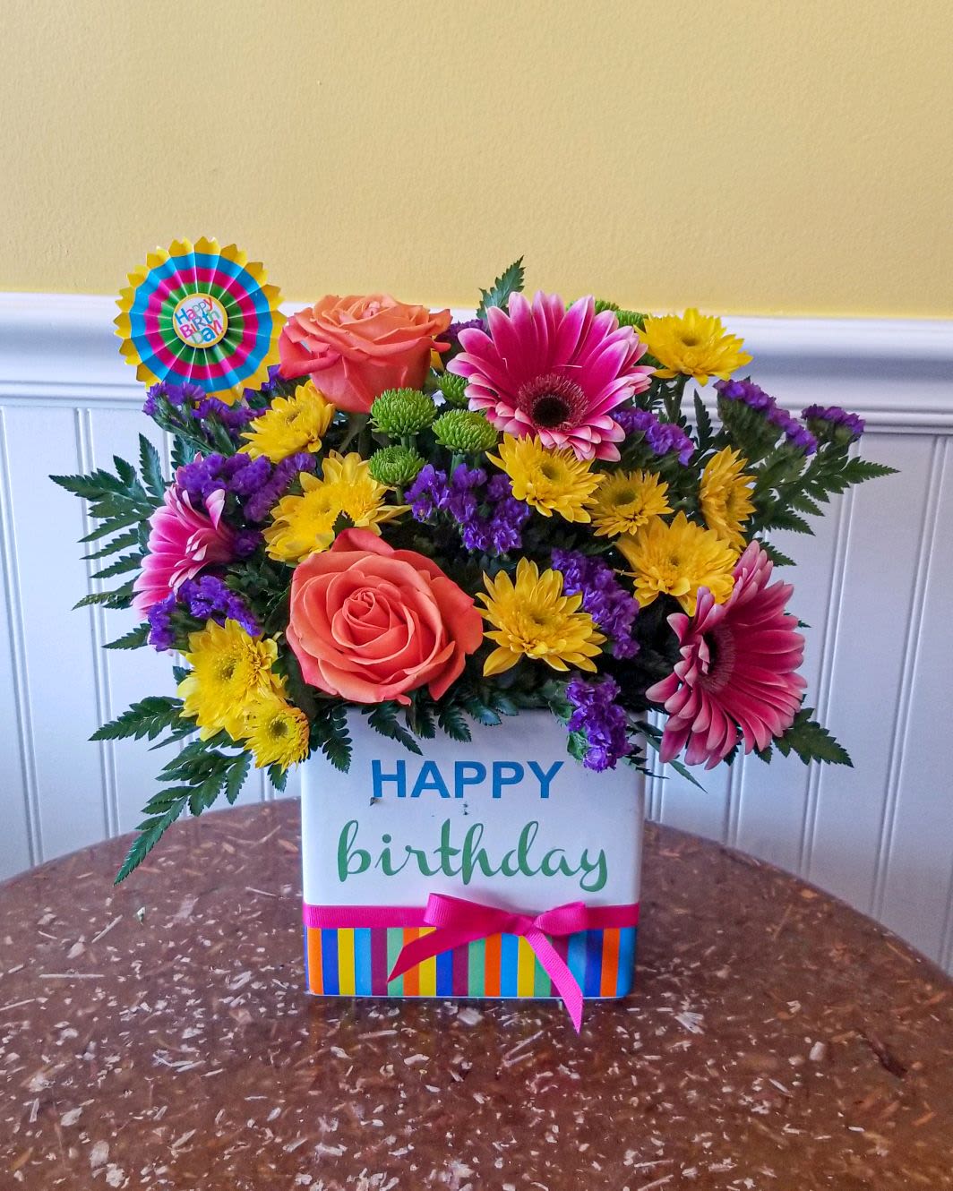  Birthday Brilliance Bouquet - Birthday Brilliance Bouquet is a true celebration of color and life to surprise and delight your special recipient on their big day! Hot pink gerbera daisies and orange roses take center stage surrounded by purple statice, yellow chrysanthemums, green button poms and lush greens to create party perfect birthday display. Presented in a modern rectangular ceramic vase with colorful striping at the bottom, "Happy Birthday" lettering at the top, and a bright pink bow at the center, this unforgettable fresh flower arrangement is then accented with a striped happy birthday pick to create a fun and festive gift. Deluxe bouquet is approx. 17"H x 13"W.