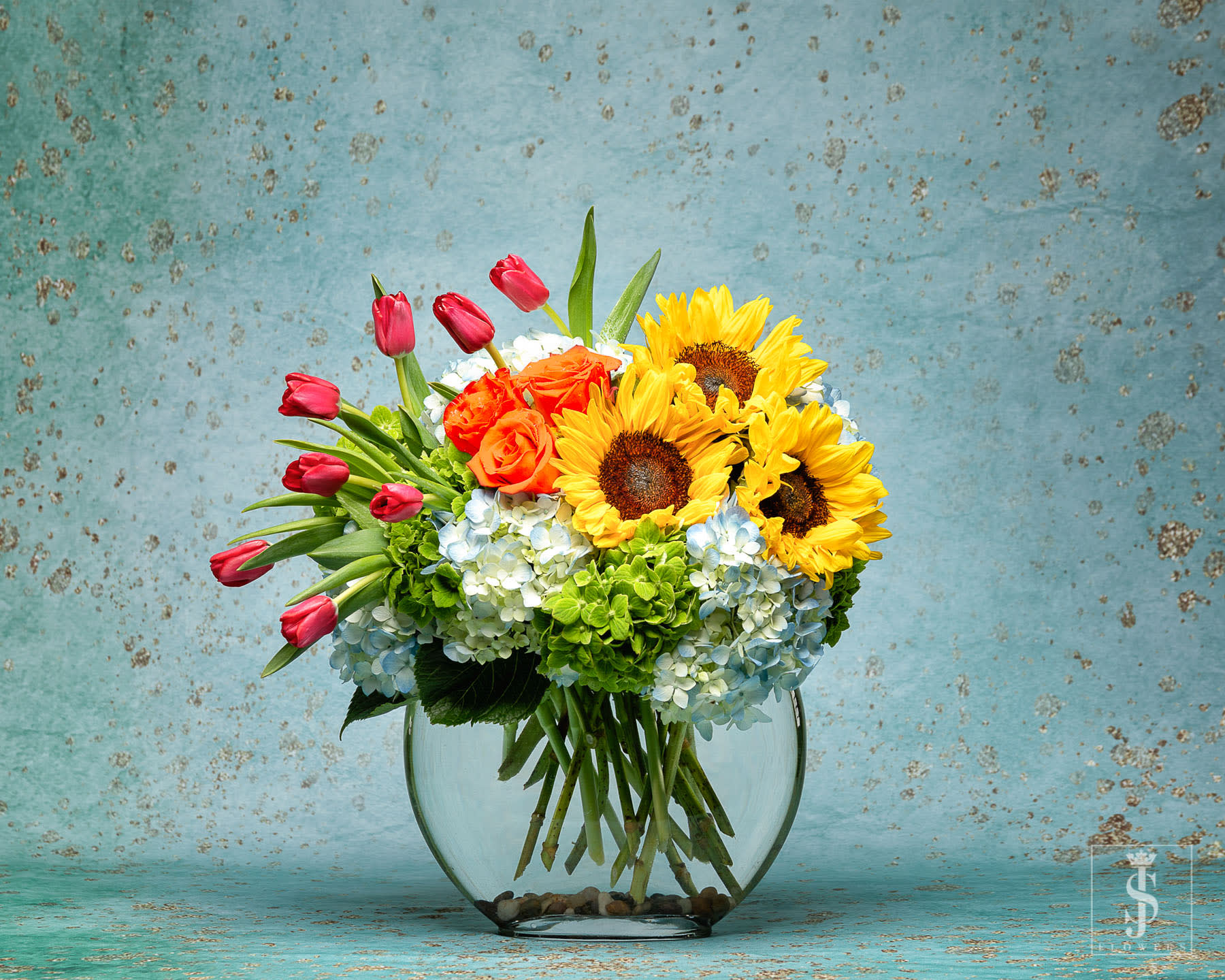Sunflower Sunset  - This beautiful arrangement captures the bright colors of Santorini's blue ocean and white hilltop houses in the glass oval-shaped modern vase, and the perfect sunset in it's lush floral selection. Yellow Sunflowers are placed amongst lush Blue and Green hydrangea, Orange Crush roses, with Tulips Falling as the sun sets.  