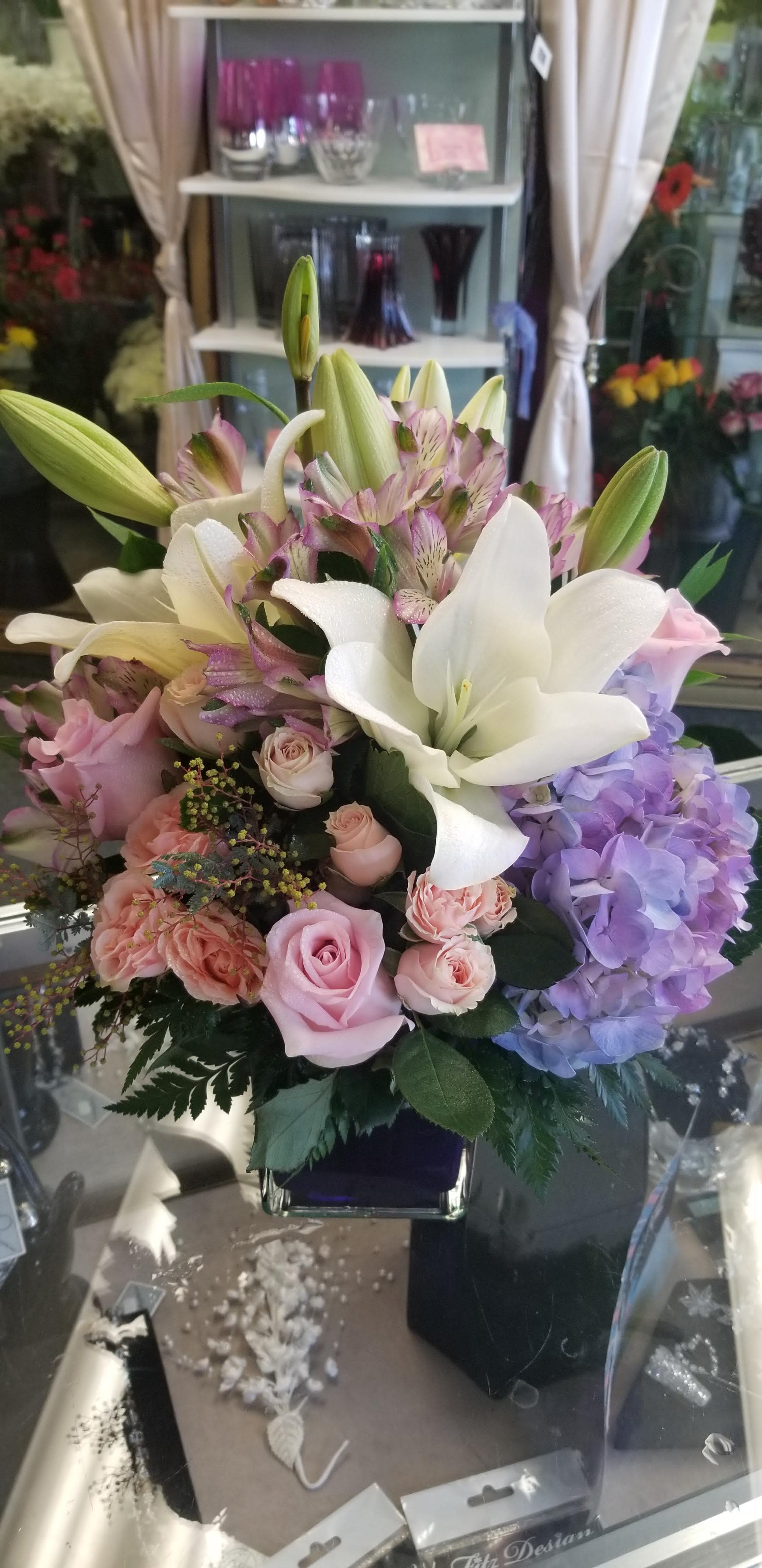 Secret Getaway by MG Florist - Designed in a modern glass cube with lilies, hydrangea, roses, spray roses, alstroemeria, and mixed greenery.  Colors may vary.