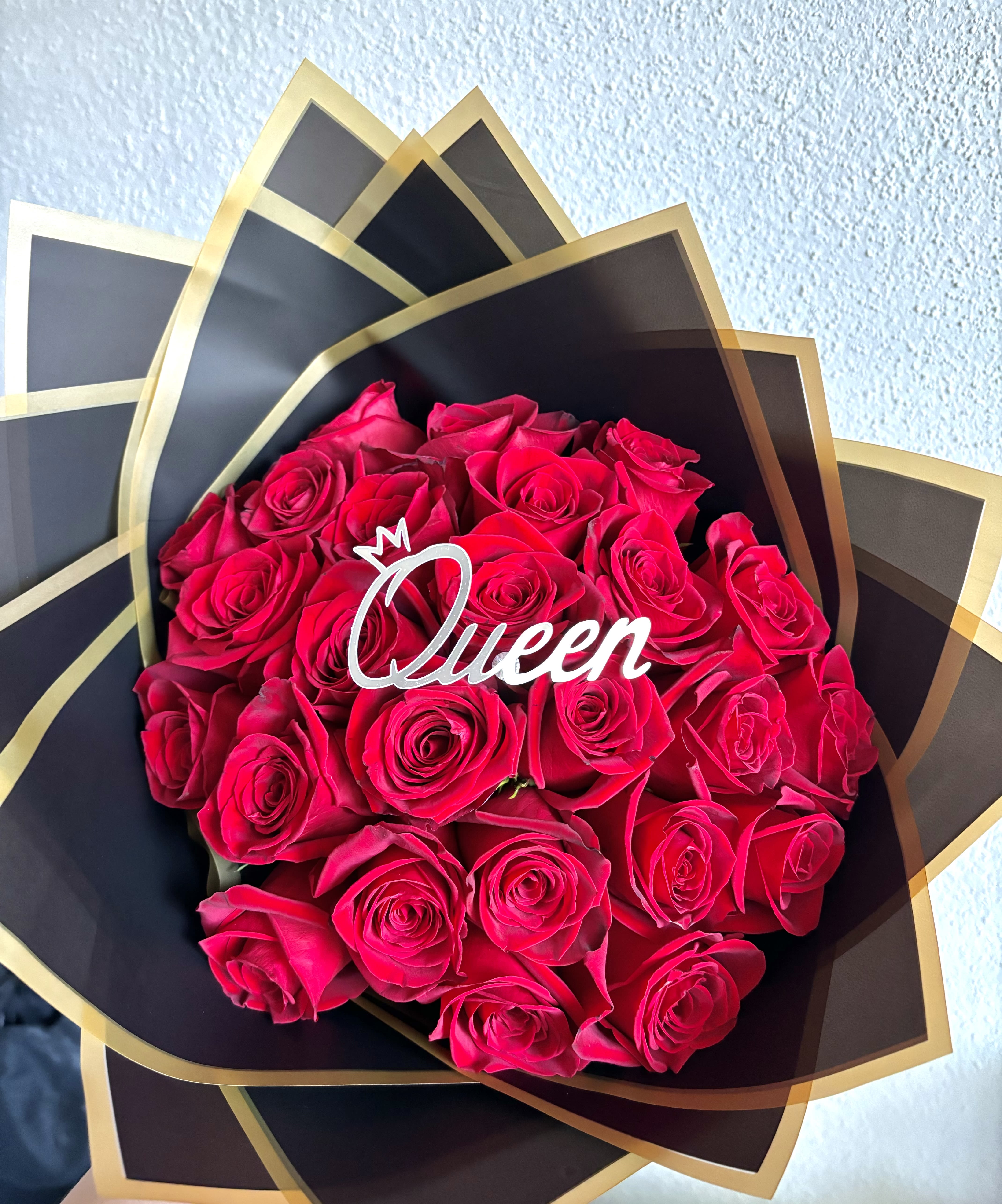 Queen Topper 25 Red Rose Bouquet - “Queen” Signature Rose Bouquet  Command attention and admiration with our elegant “Queen” Signature Rose Bouquet — a radiant arrangement of velvety red roses, wrapped in sleek black and gold for a truly majestic presentation. Each petal speaks of passion, strength, and grace, making this bouquet a stunning tribute to the queen in your life. Topped with a chic “Queen” plaque, this design is perfect for birthdays, anniversaries, or simply to remind her of the royalty she is. Elevate your floral gifting with timeless beauty and regal flair. 