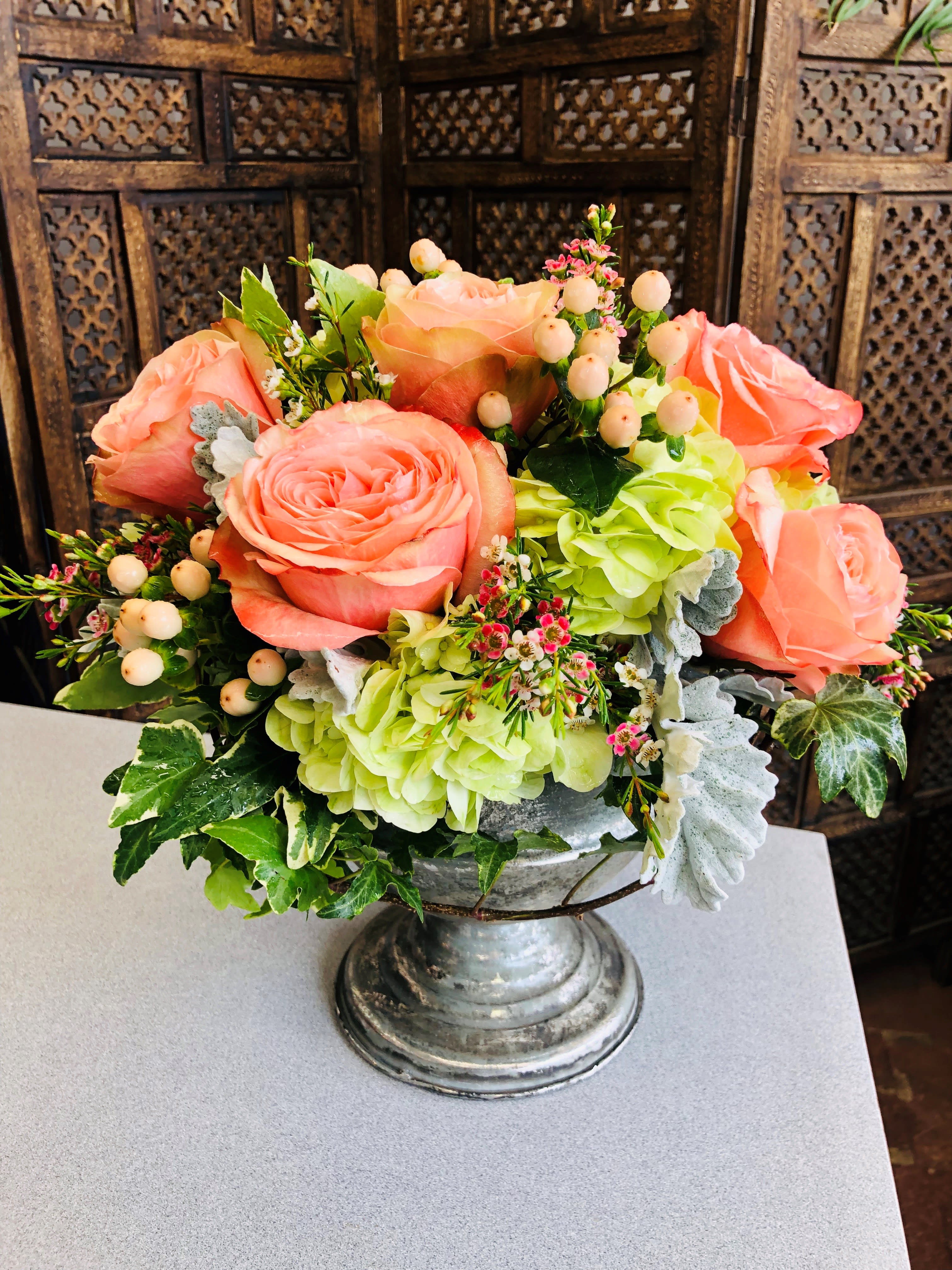 Kahala Dreams - This Floral Design is highlighted by beautiful Peach roses set against a back ground of Hydrangea ans complimented with Hypercium AKA Coffee Bean.  Created in a metallic urn.