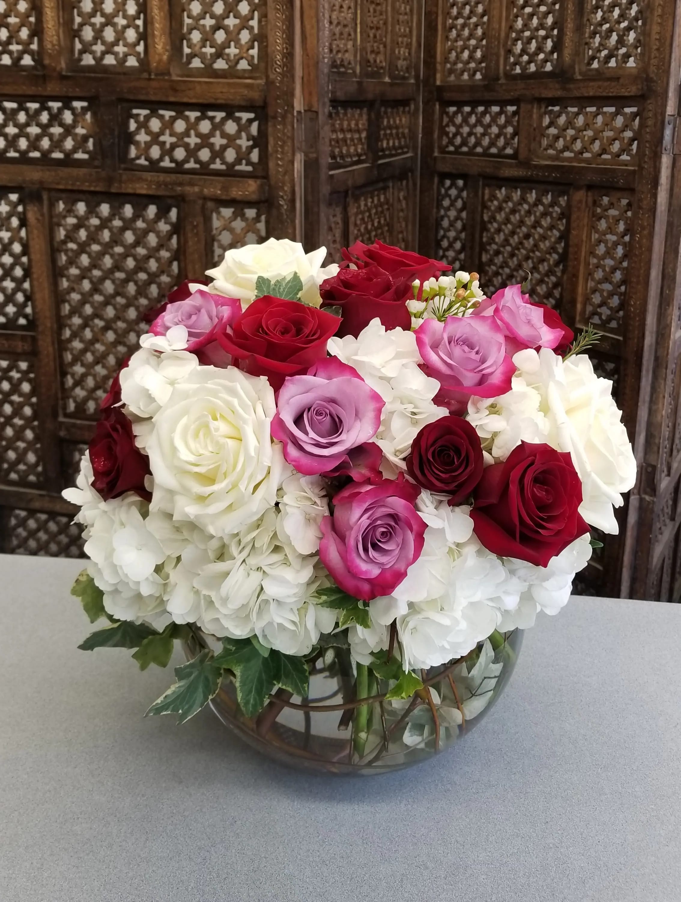 Looking Good - Looks can be deceiving.  This large sphere is filled with 24 roses mixing Deep Purple, Red, and White. Atop a cloud of White Hydrangea with other small flowers and greenery.  Inside the Vase is an array of Curly Willow bringing interest inside the vase.