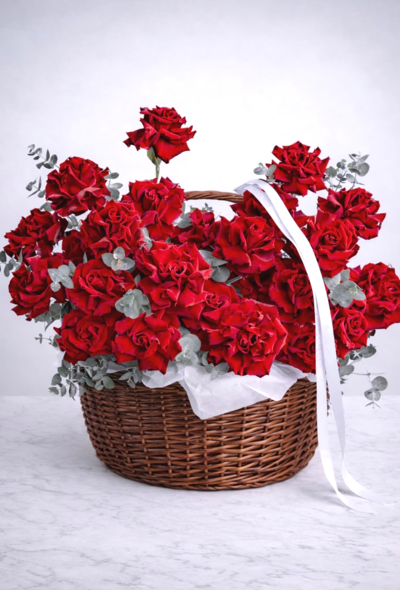 Rustic Romance Rose Basket - A stunning celebration of classic romance and natural elegance — the Rustic Romance Rose Basket features a lush bouquet of premium long-stem red roses artfully arranged with soft silver eucalyptus and presented in a beautifully crafted natural brown wicker basket. Finished with delicate white tissue and a flowing satin ribbon, this arrangement blends timeless style with a warm, rustic charm — perfect for anniversaries, Valentine’s Day, weddings, or special moments worth remembering.