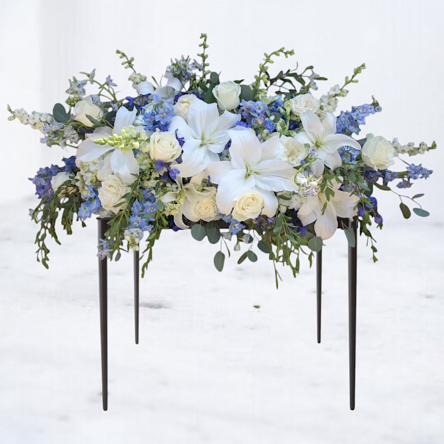 Reverence Veil Casket Spray - Ethereal and serene, the Reverence Veil casket spray is a lush tribute designed to honor a loved one with grace and dignity. Featuring a soothing blend of ivory lilies, creamy white roses, pale blue delphinium, and soft cascading greens, this arrangement evokes a sense of peace and gentle remembrance.  Its flowing composition drapes beautifully over the casket, offering a quiet yet profound expression of farewell. Ideal for those seeking a refined and heartfelt way to celebrate a life filled with love, light, and legacy.