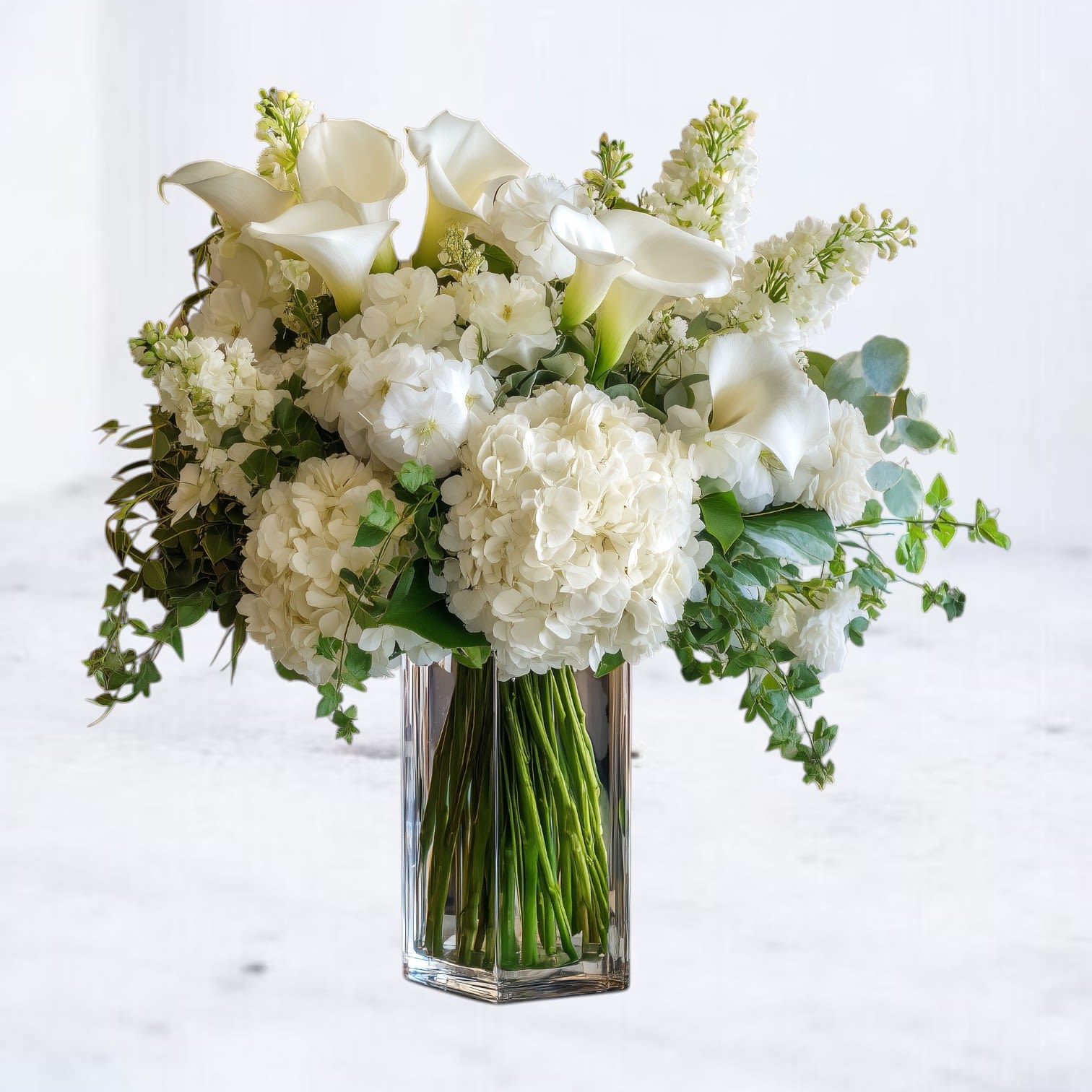 Whitehall - Whitehall is a sculptural all-white arrangement designed with balance and quiet impact. Featuring lush hydrangea, elegant calla lilies, and seasonal white blooms accented with natural greenery, this composition highlights texture and form in a clean, modern silhouette.  Arranged in a clear glass vase, Whitehall offers a refined presence that complements contemporary interiors and elevated spaces. Its monochromatic palette makes it a versatile choice for celebrations, milestone moments, corporate gifting, or simply bringing understated luxury into the everyday.  Available for same-day flower delivery in Magnolia, The Woodlands, Spring, Conroe, and throughout the Greater Houston area. Flower varieties may vary seasonally while preserving the overall style, fullness, and structure of the design.