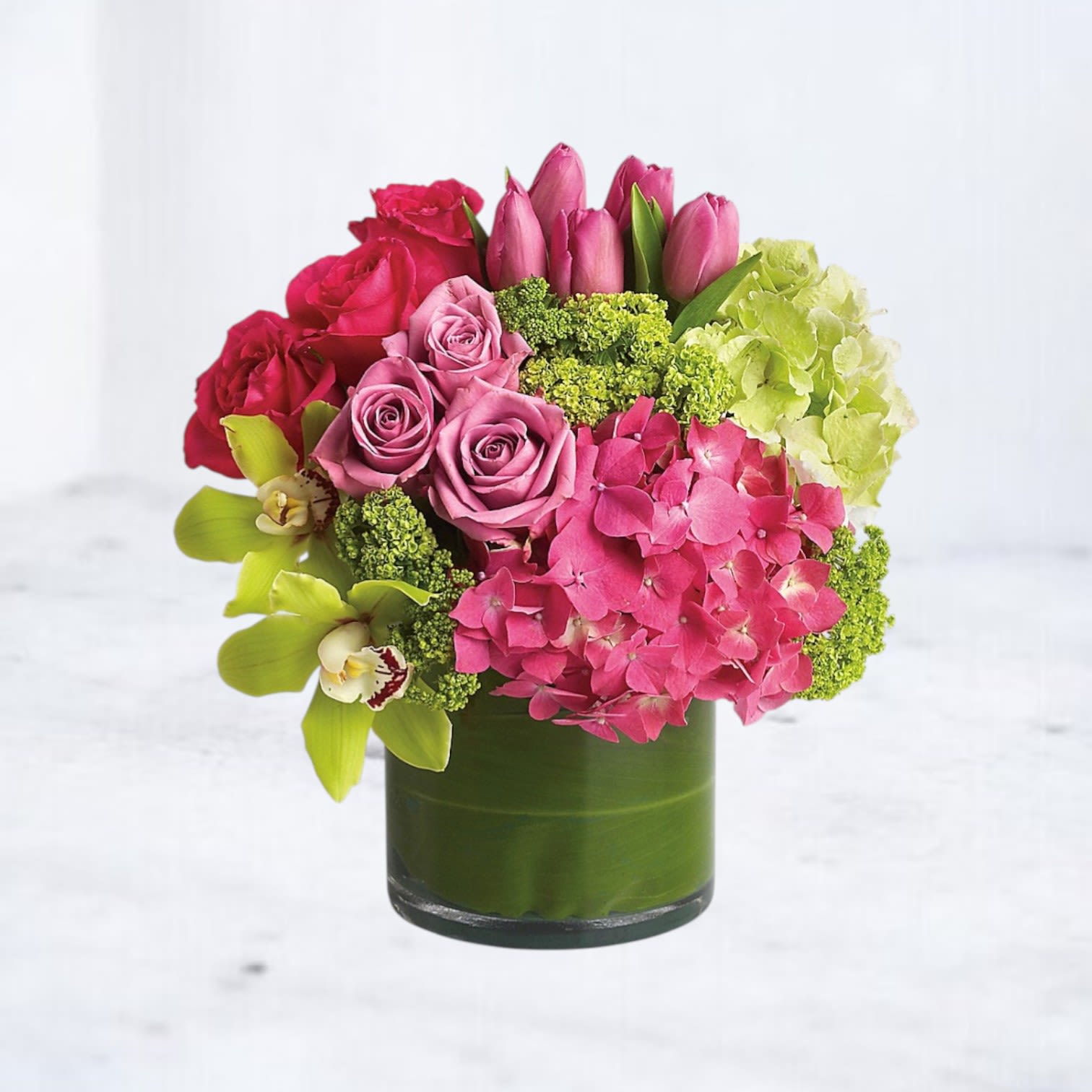 Blushing Orchid Delight - Add a touch of elegance with this stunning floral arrangement, featuring a blend of pink roses, tulips, hydrangeas, and exotic green orchids. Nestled in a chic glass vase wrapped with a decorative green leaf, this bouquet is perfect for brightening up any space, celebrating special occasions, or expressing love and gratitude. With its lush mix of fresh flowers and vibrant colors, this arrangement makes an ideal gift for birthdays, anniversaries, or just because. Order this exquisite flower bouquet to bring joy and beauty to someone’s day.  Disclaimer: In the event the orchids shown in the photo are not available, another variety of orchids will be used to maintain the beauty and quality of the arrangement.