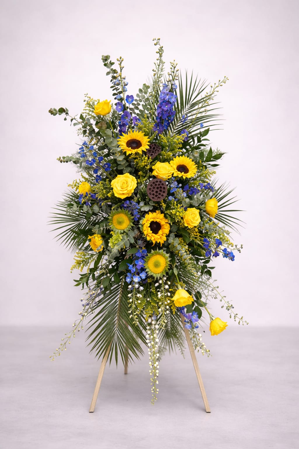 Ray of Hope Standing Spray - Bring warmth and comfort to a time of loss with our Ray of Hope standing spray. Golden sunflowers and radiant yellow roses shine brightly against calming shades of blue delphinium and lush greenery, creating a heartfelt tribute that reflects both love and remembrance. The uplifting color palette symbolizes light breaking through sorrow — a reminder of cherished memories and brighter days ahead.  Displayed on a standing easel, this spray is a beautiful way to honor a life well lived and to surround loved ones with hope and peace. Custom color selections are available upon request to create a truly personal expression of sympathy.