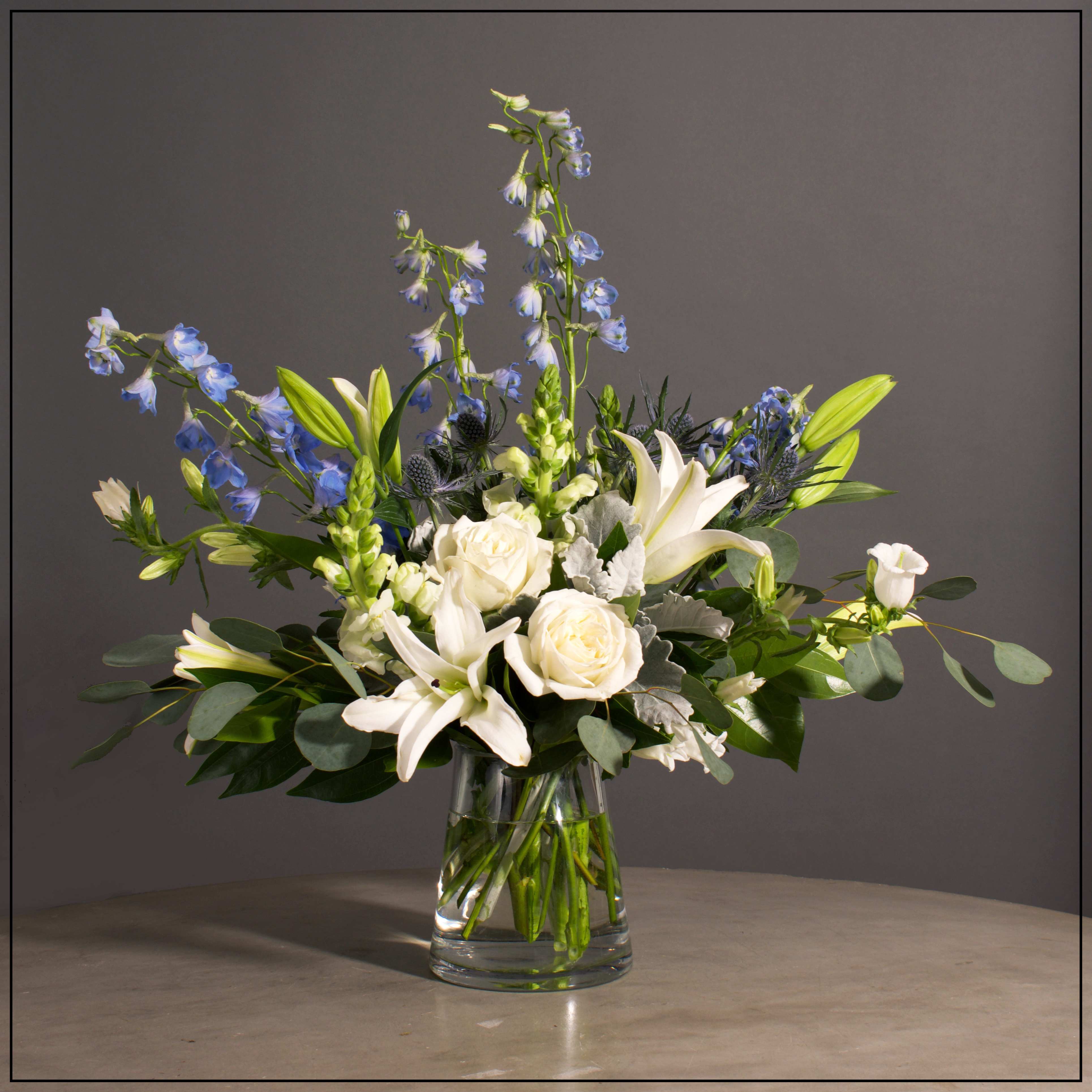 Brooklyn - Delphinium, Lillies, Thistle, Campanula and Snapdragons with Premium Greens in a unique Glass Vase.  Approximately 23" H x 28" W