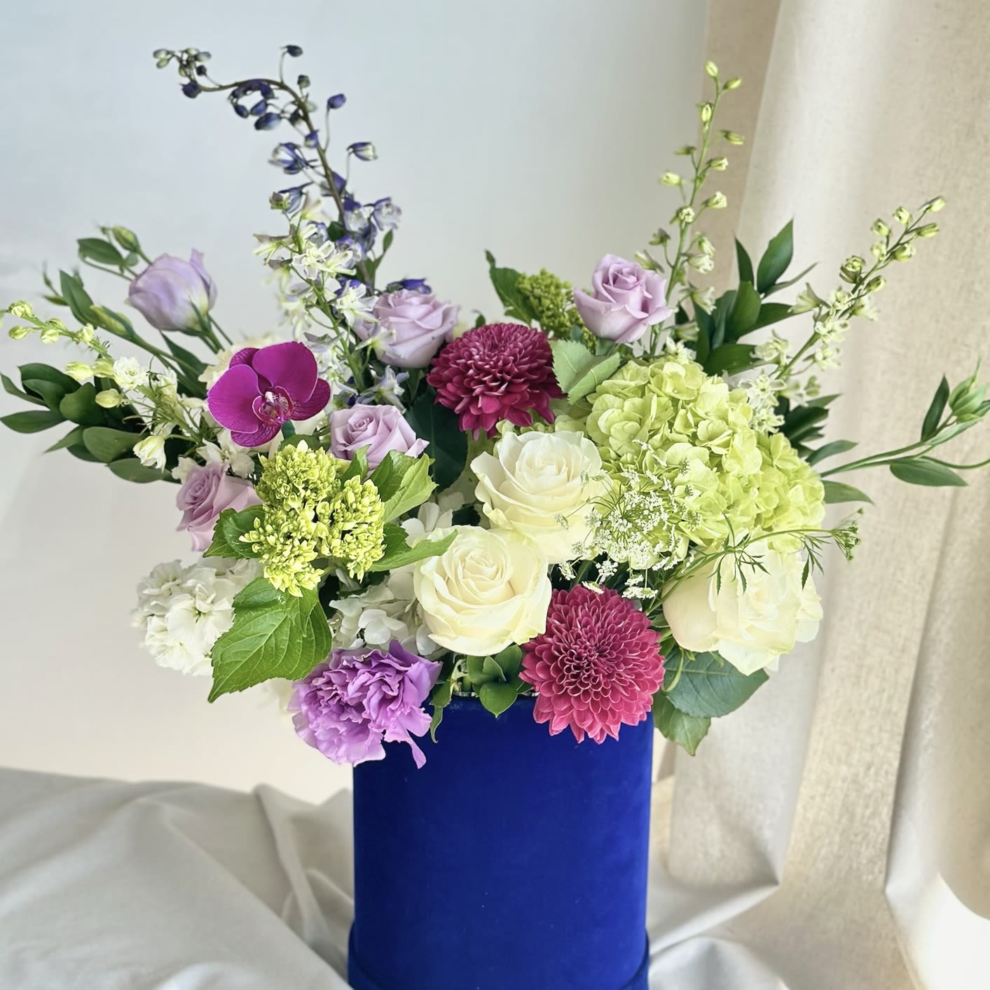 Blue Violet  - A tall round velvet blue box filled with a mix of white, blue, purple, green, and lavender flowers.   Flower types may vary depending on the season.