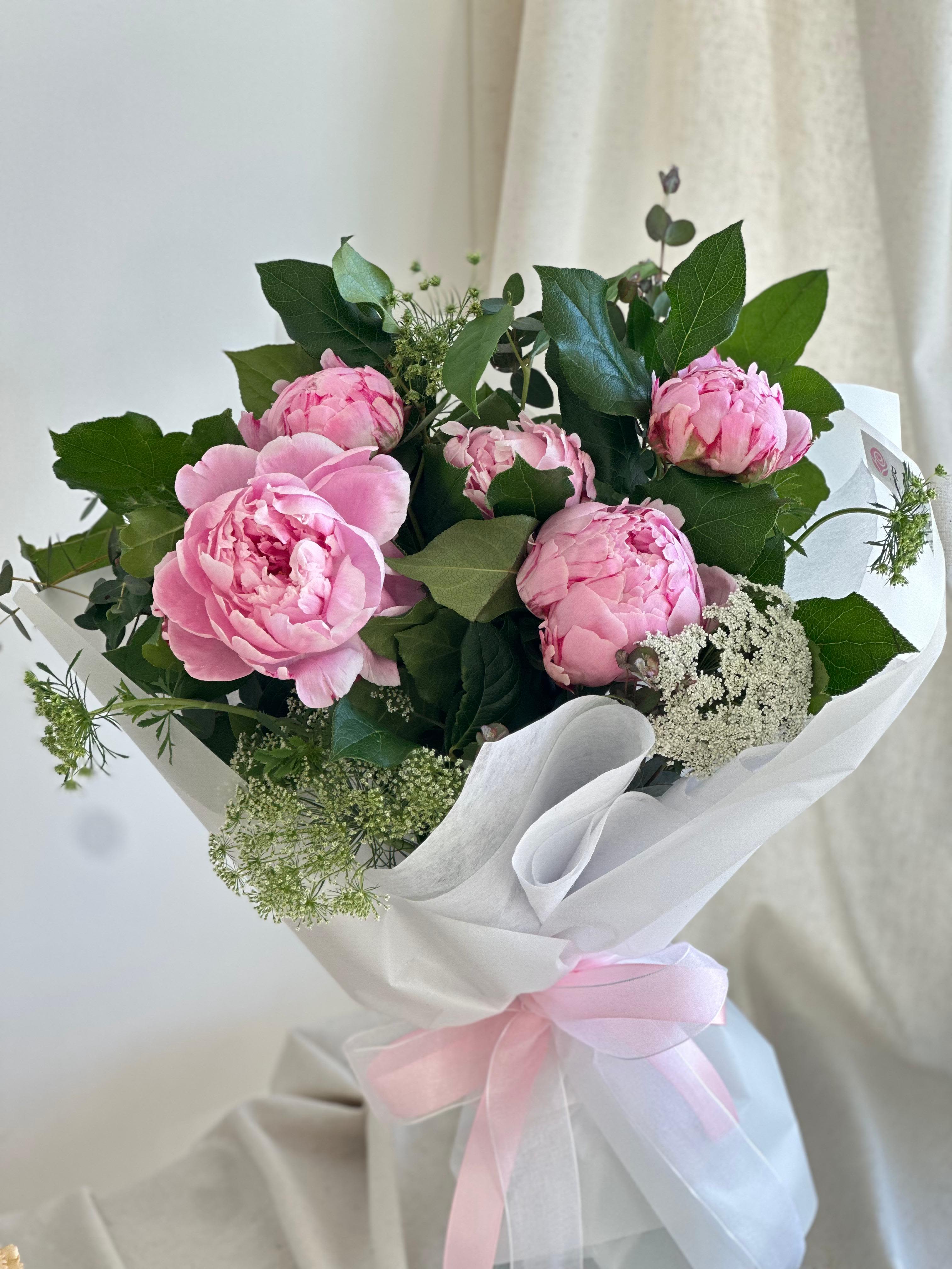 Peonias in Rosa - A round bouquet of peonies with plenty of greenery and fillers, wrapped in white paper.   Flower types may vary depending on the season.