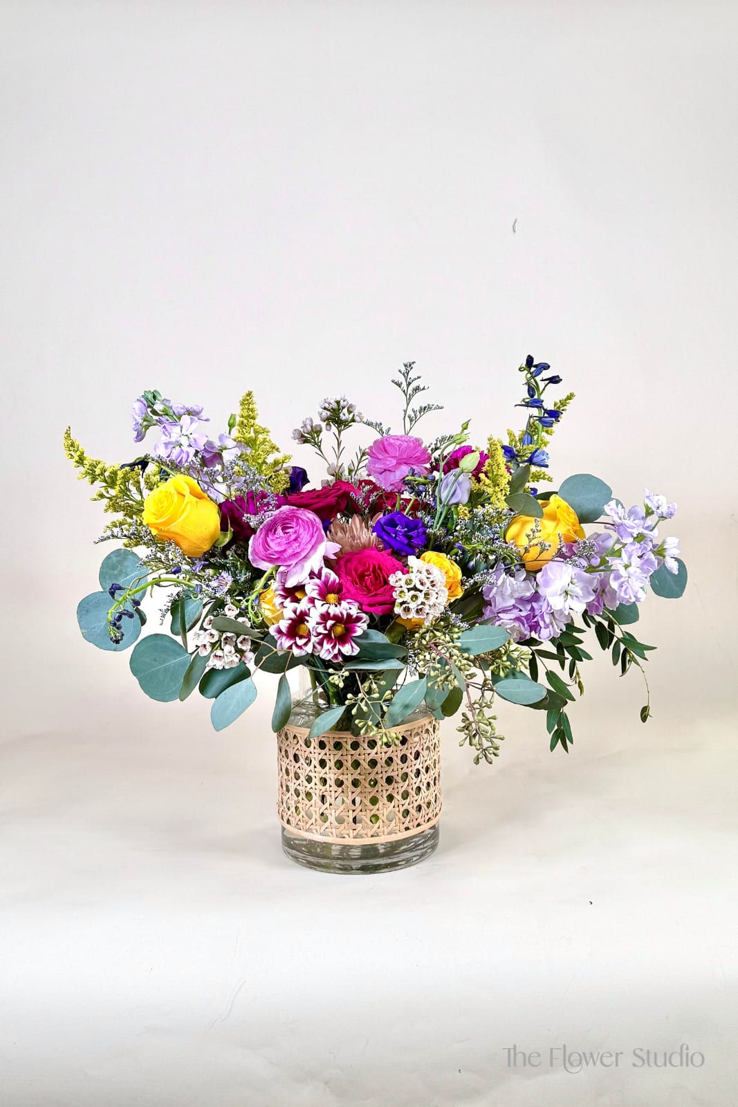 Monets - Bright and cheerful, this garden-inspired arrangement bursts with vibrant yellows and striking blues. Featuring golden roses, gerbera daisies, and seasonal blooms paired with lush greenery, it’s beautifully designed in a clear glass vase with a woven accent wrap. Perfect for birthdays, celebrations, or simply bringing a touch of sunshine indoors, this arrangement radiates happiness and charm. Approx. 15" H x 20" W Our floral artists hand-design each arrangement, so flower and container styles may vary based on local and seasonal availability. As a result, we are unable to guarantee specific florals, colors, or vessels—but we always ensure the same level of beauty and value in every piece.