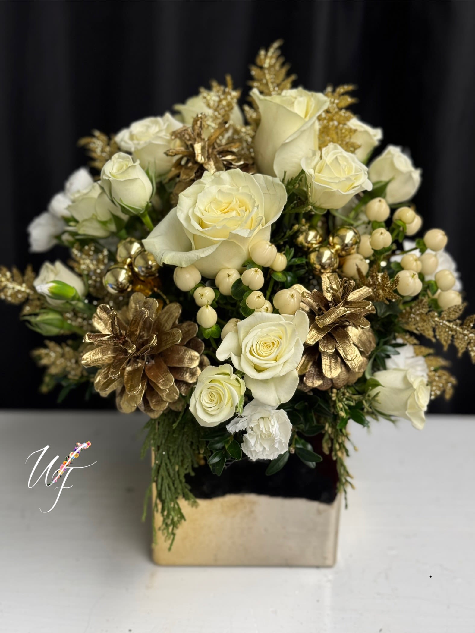 White roses in a gold-toned square vase with gold pinecones