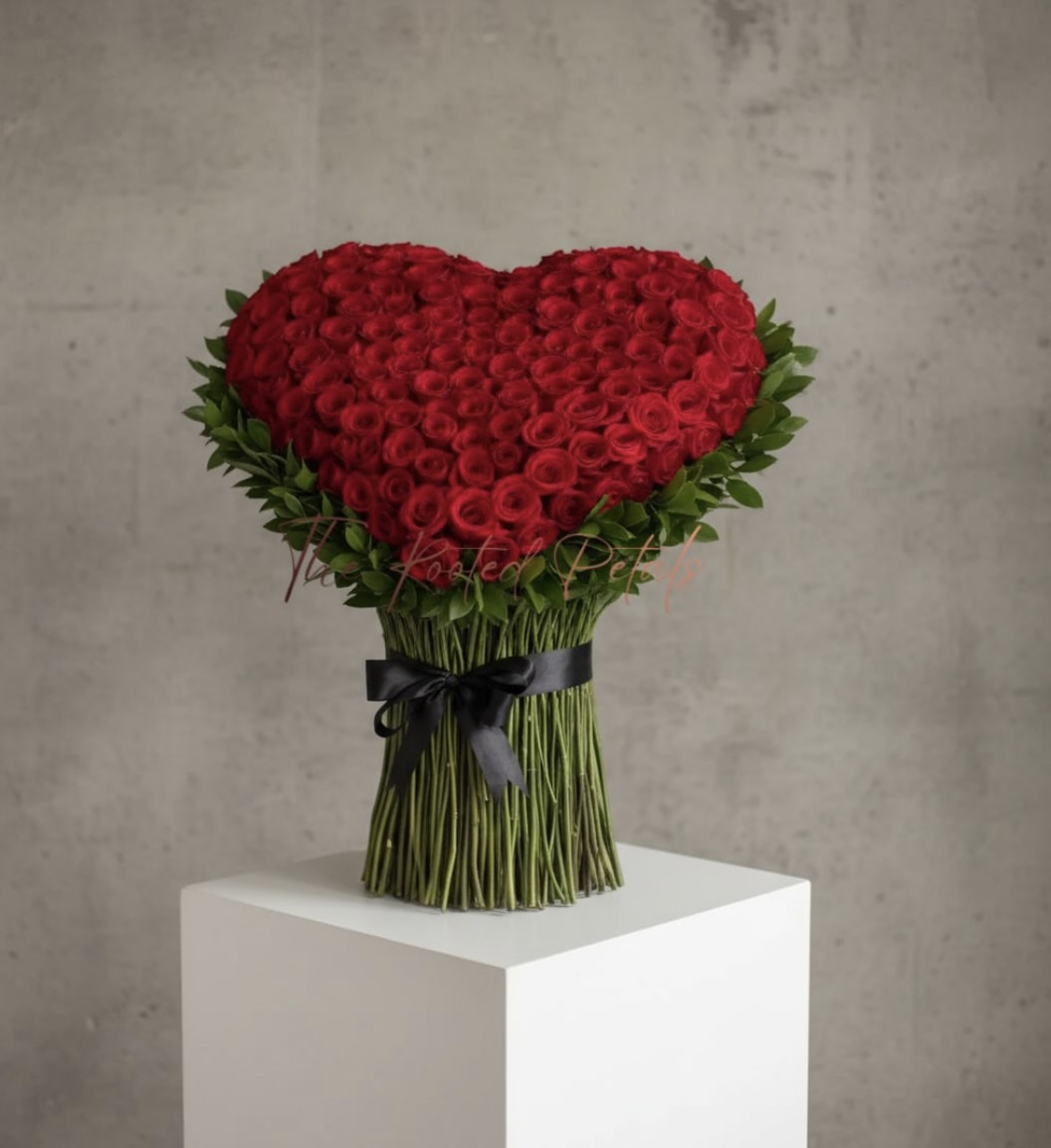 Standing Blooming Heart - Make a statement with the Standing Blooming Heart — a sculptural bouquet of tightly clustered crimson roses arranged into a perfect heart and mounted upright on a neat, hand-tied stem pedestal. Glossy green foliage frames the heart’s edge for fresh contrast, while a black satin bow at the base gives the arrangement a modern, refined finish. 