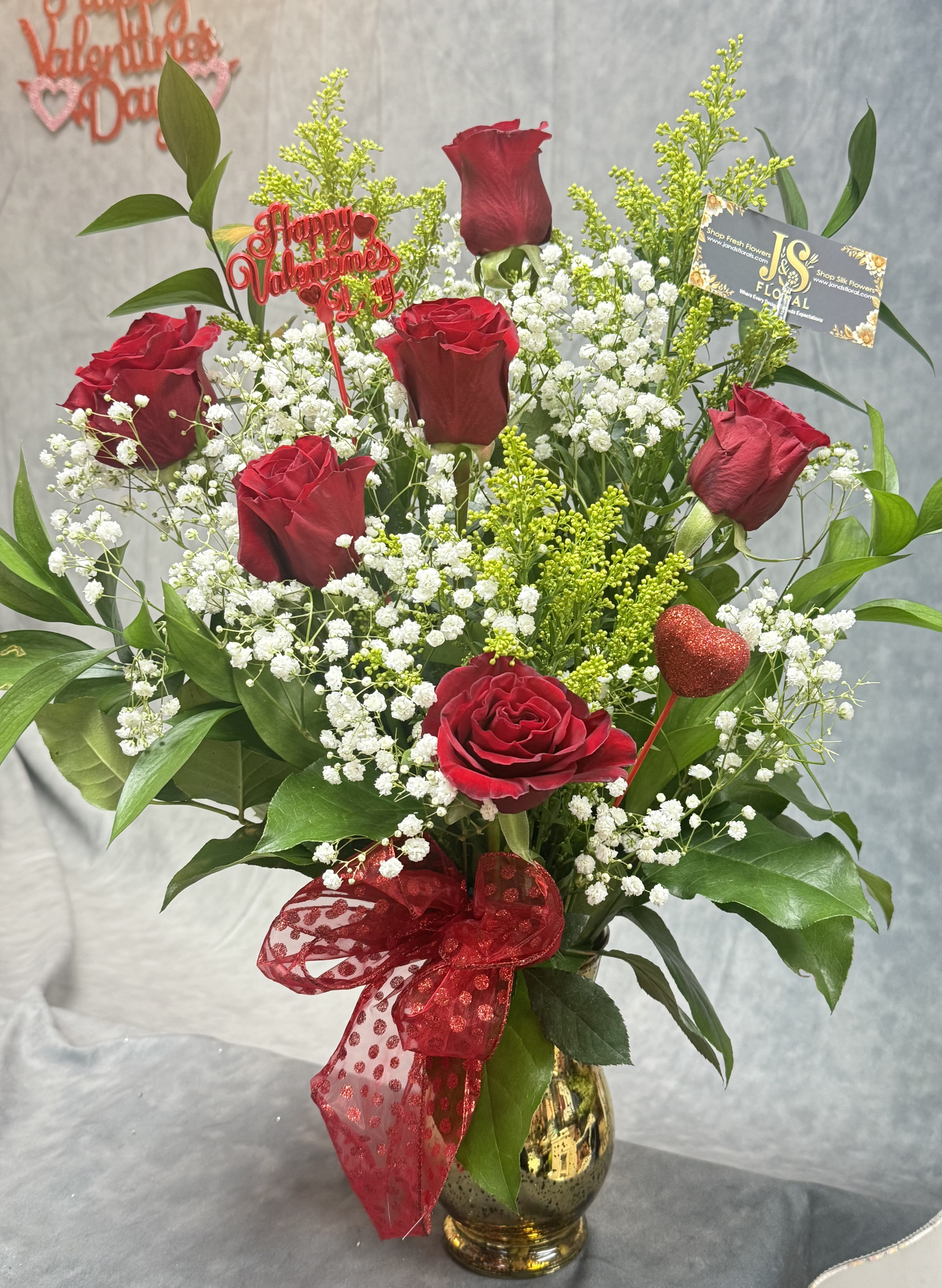 Red Roses Front Face Vase - A charming mixed-rose arrangement featuring red roses accented with fresh greenery and golden filler, elegantly styled in a vase and finished with a red ribbon—perfect for expressing love, appreciation, or a sweet everyday surprise. 
