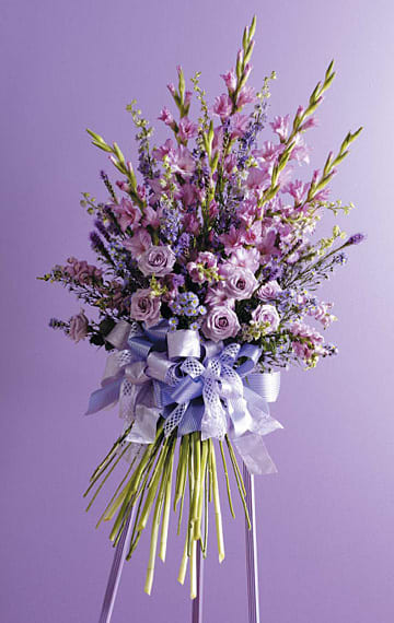 Lavender Peace Standing Spray - Soft and soothing, the Lavender Peace Standing Spray offers a graceful expression of sympathy and remembrance. Designed in calming shades of lavender and violet, this arrangement features elegant gladiolus, lush roses, stock, and limonium, all accented with delicate greenery and finished with a beautifully layered ribbon. Its tranquil palette and airy structure make it a heartfelt tribute to honor a loved one’s memory with gentleness and grace. What’s included? Lavender Gladiolus, Lavender Roses, Stock, Limonium, and Seasonal Foliage Finished with a decorative bow and presented on a standing easel for service display.