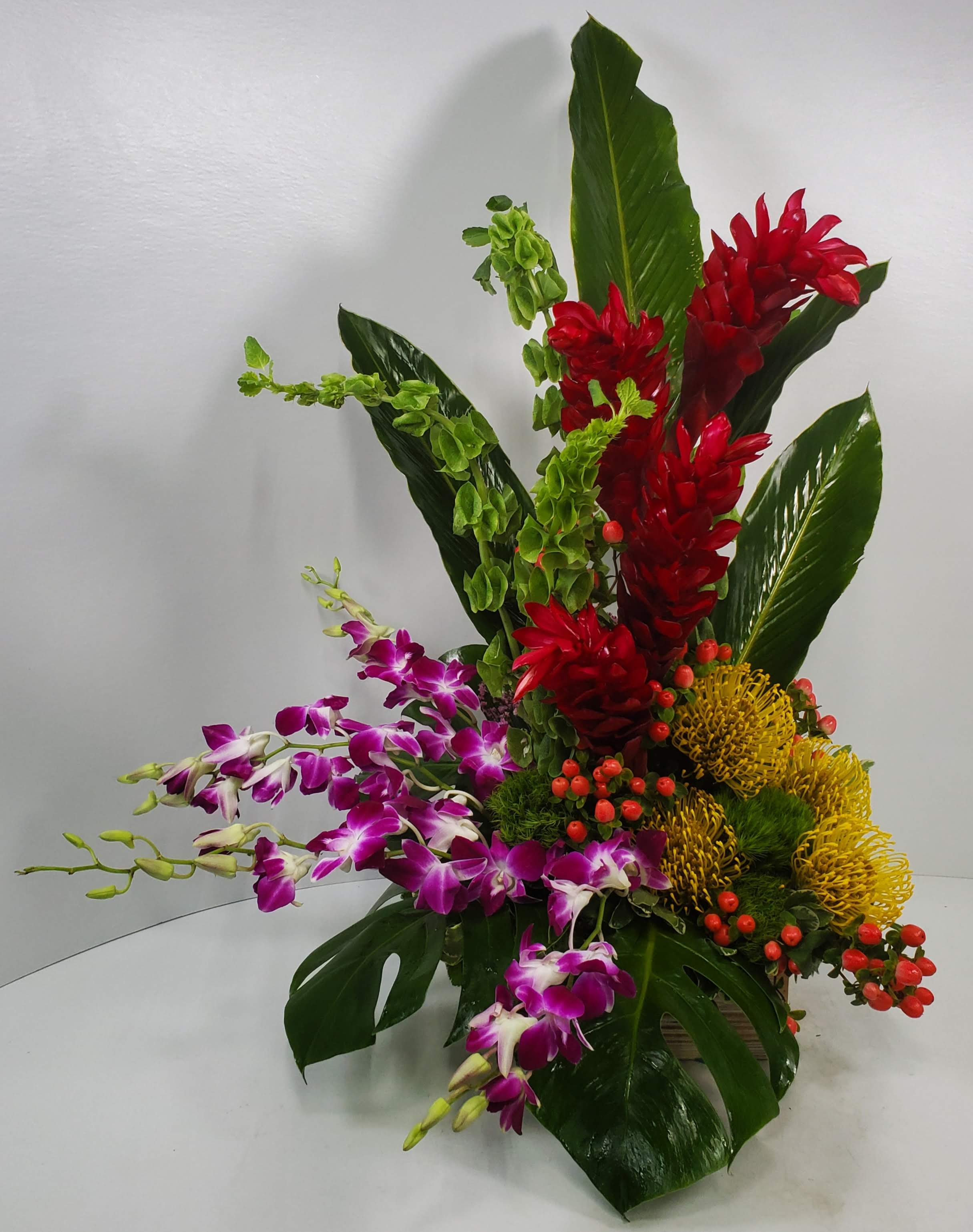 TROPICAL CELEBRATION - Make a bold tropical statement with this striking vertical arrangement featuring dramatic red ginger, golden pincushion proteas, cascading purple orchids, and lush greenery. Accented with bright berries and layered foliage; it delivers vibrant color and rich texture. This arrangement is perfect for celebrations, grand openings, or anyone who loves a modern island-inspired design.