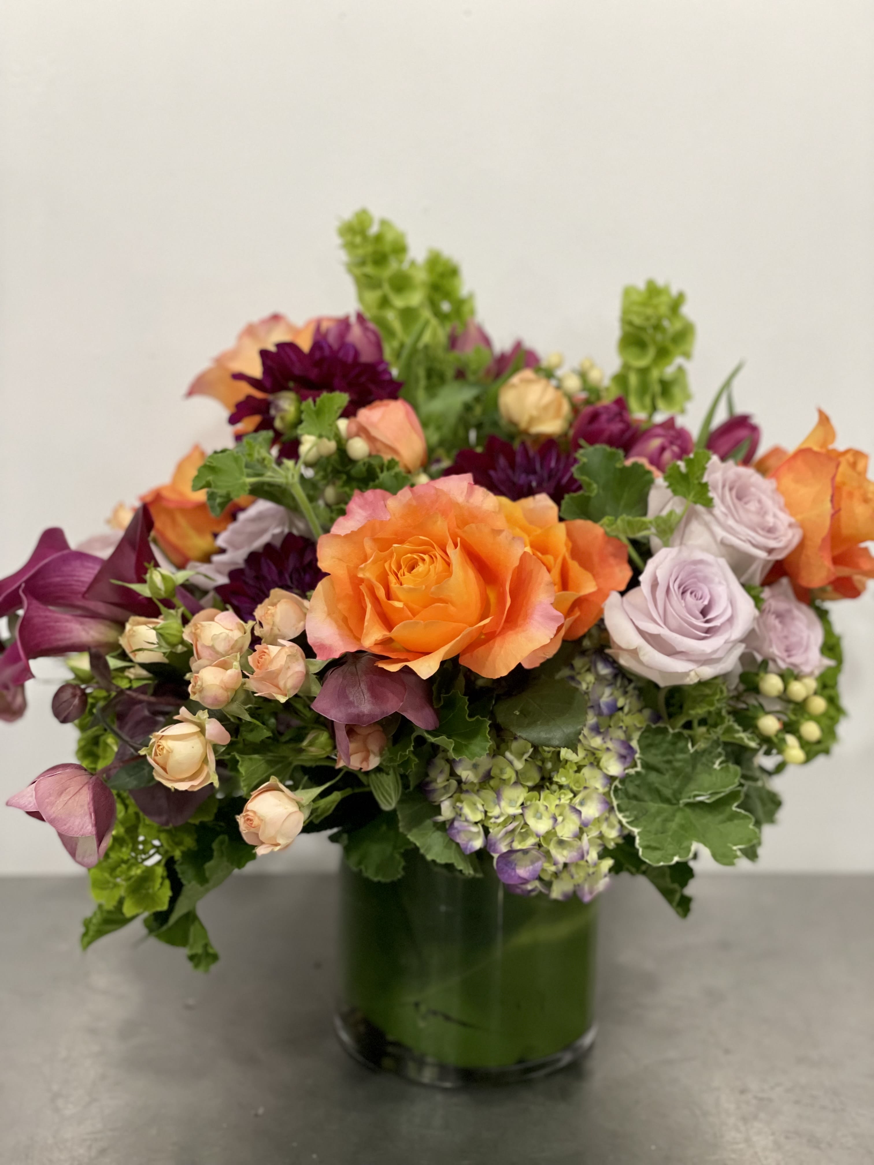Summer lovin' - All the ingredients are seasonal and bound to create an impactful piece for any room. This one in particular has been popular for its color palette designed in a compact, luxe way. This arrangement features premium blooms in purples, lavender, peach, and orange with bright green. Pictured here is the deluxe size.   If any design element such as color or flower/foliage are very important to your order please contact us to ensure availability first by calling the shop at  415 751 2747