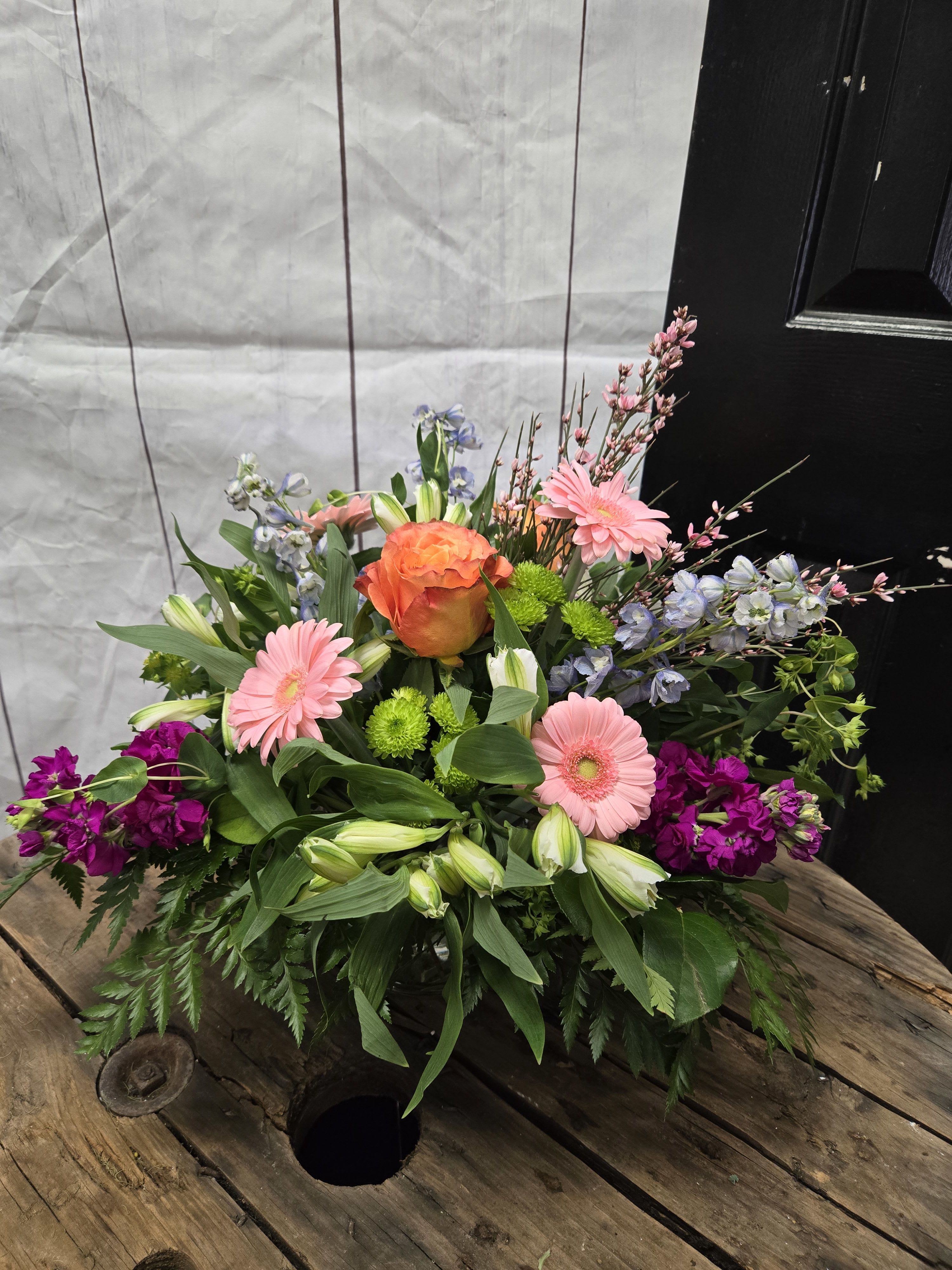  Radiance - This floral arrangement features a delightful blend of vibrant and soft tones. At its heart is a striking orange rose that radiates warmth and draws the eye. Surrounding it are pastel pink gerbera daisies, adding a touch of softness, while the deep purple stock flowers introduce a rich, elegant contrast.  Delicate blue delphiniums and bright green button mums add depth and freshness, that offer a hint of sophistication. The arrangement is rounded out with lush greenery and sprigs of pink blossoms, creating a full and balanced look. It’s a perfect display for springtime celebrations or to brighten any space with cheerful energy *All flowers are subject to substations with seasonal availability 