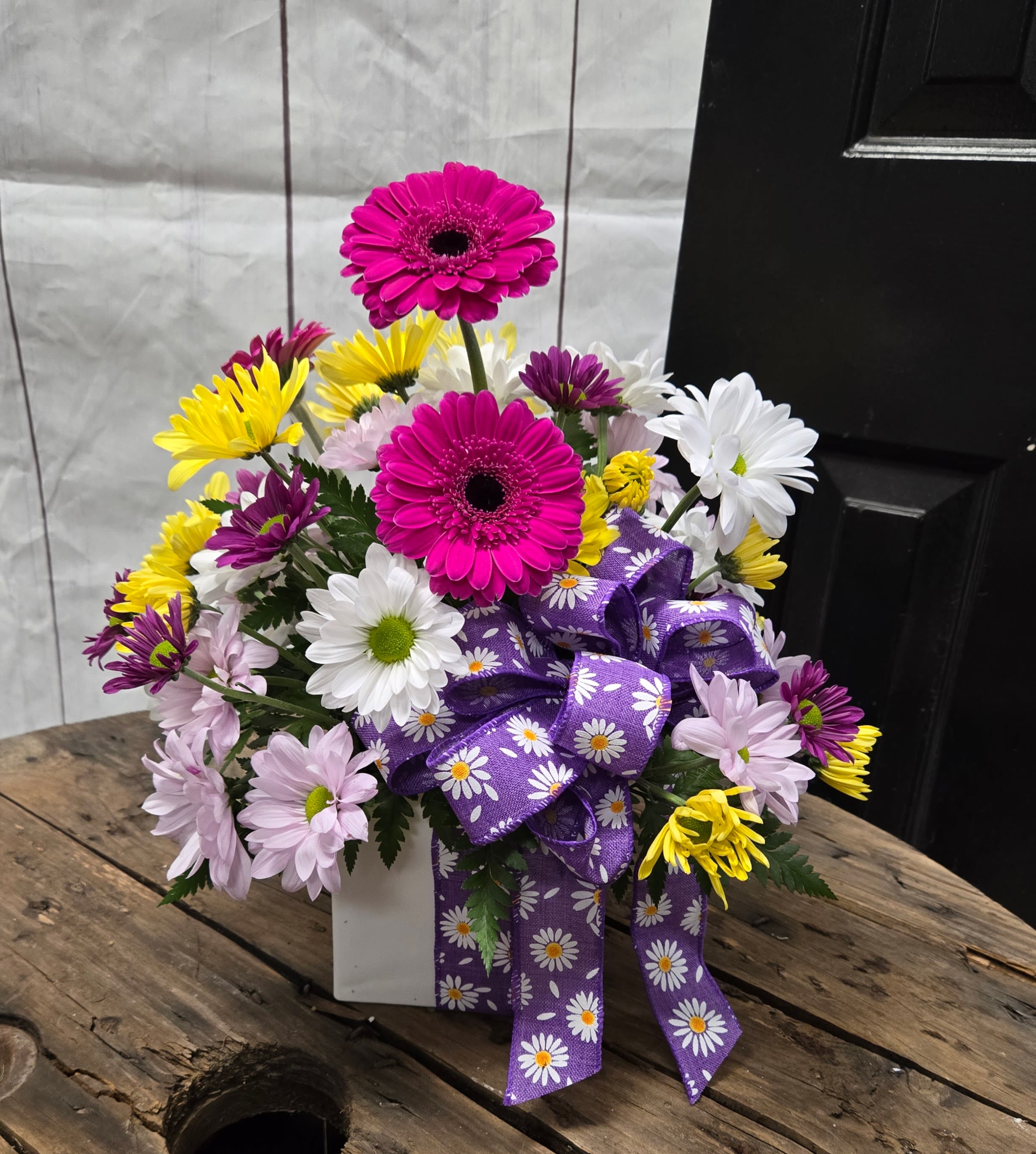 Coming up Daisies - This vibrant arrangement features a cheerful mix of colorful daisies and gerbera daisies in shades of hot pink, white, yellow, and light purple. The playful design is accented with a purple ribbon adorned with daisy patterns, adding a touch of charm and whimsy. The flowers are arranged in a white rectangular container, creating a bright and joyful centerpiece suitable for various occasions, from celebrations to thoughtful gifts.  * All flowers are subject to substations with seasonal availability