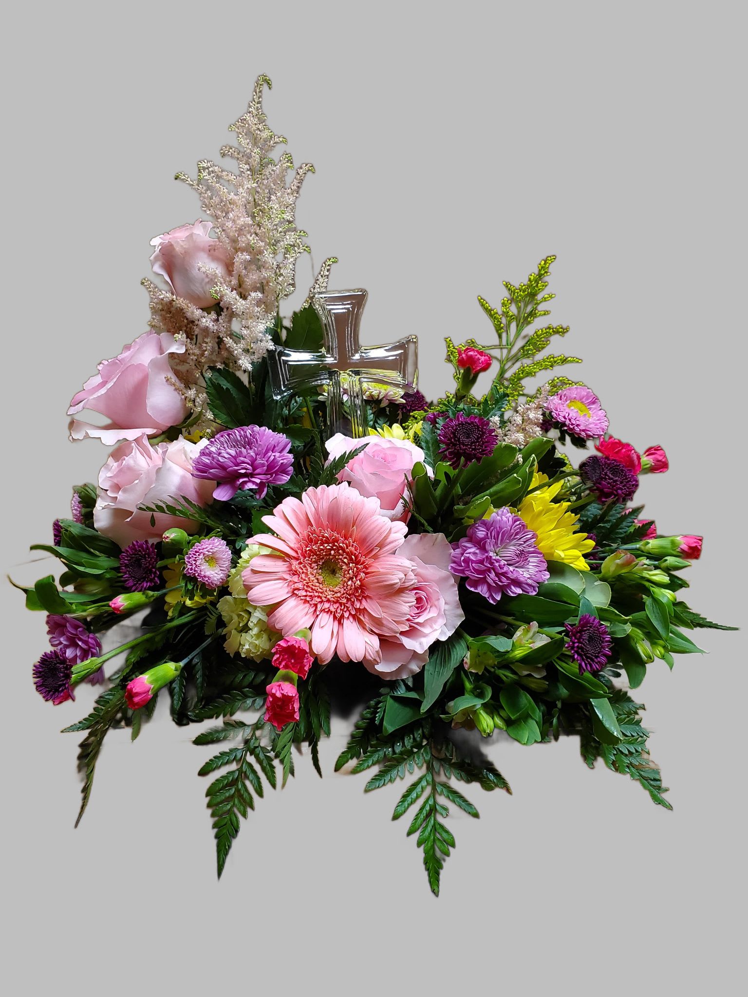 Graceful Remembrance - This floral arrangement exudes elegance and tranquility, making it a fitting centerpiece or tribute piece.   Soft Pink Roses: Adding a touch of romance and grace, symbolizing love and admiration. Pink Gerbera Daisies: These cheerful blooms bring vibrancy and warmth, representing innocence and purity. Purple Chrysanthemums: A pop of richness, offering depth and a sense of reverence. Yellow Accents (likely sunflowers or similar): Brightening the arrangement with positivity and hope. Greenery: Including ferns and other foliage, providing a lush backdrop and grounding the design. Glass Cross Ornament: A central element symbolizing faith, spirituality, and peace. This arrangement is perfect for a memorial service or religious occasion, combining a harmonious blend of color, texture, and sentimentality. * All flowers are subject to substations with seasonal availability.