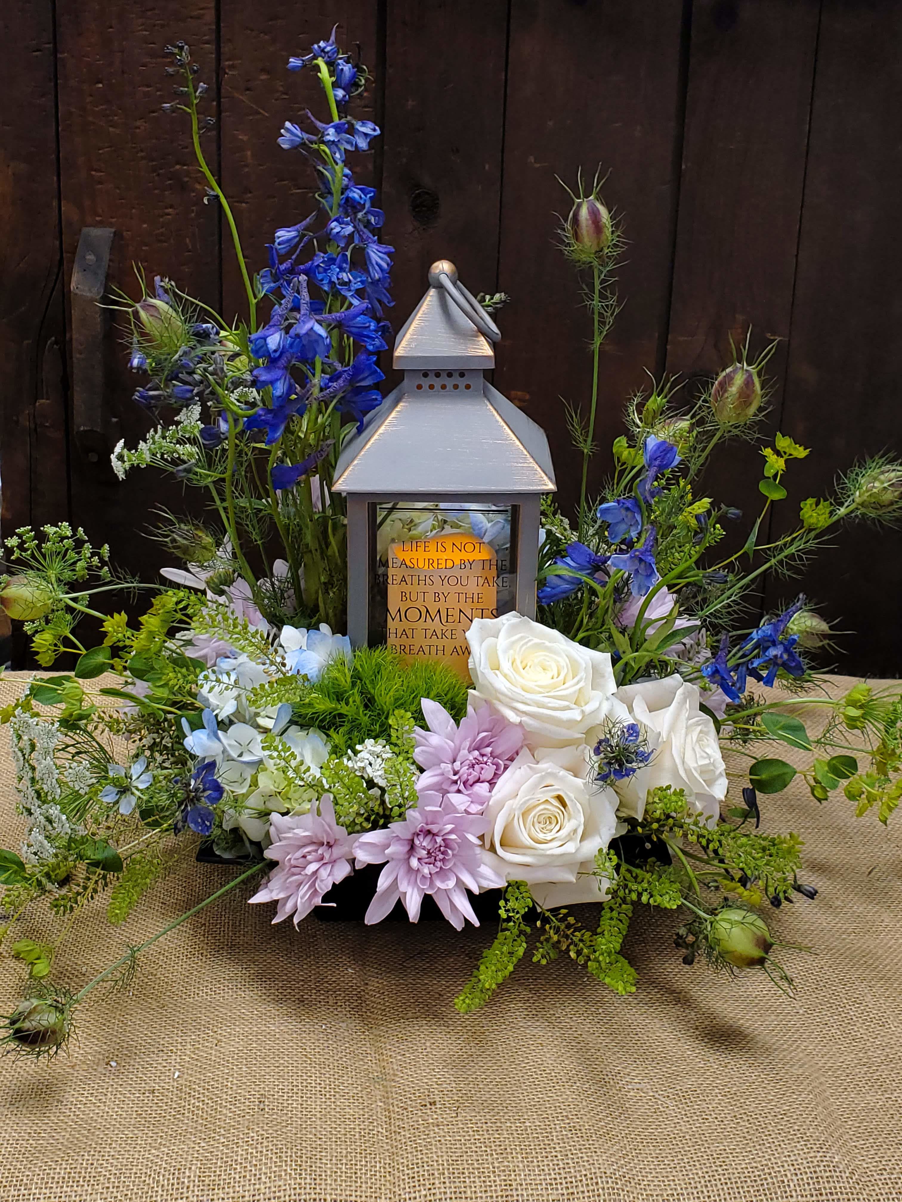 Lovely light  -  This arrangement is a stunning and serene design that incorporates both floral and decorative elements to create a meaningful centerpiece.   Centerpiece Feature: A lantern with a heartwarming message,  serves as the focal point, adding a sentimental touch to the design. Saying on lanterns may vary. Floral Details: Blue delphiniums create height and provide a vibrant pop of color, drawing attention upward. White roses add elegance and purity, balancing the vibrant tones with their classic beauty. Light purple chrysanthemums offer a soft, pastel hue that complements the arrangement. Greenery and fillers, such as Queen Anne's lace, nigella pods, and moss-like textures, contribute to the natural and whimsical feel, adding depth and volume. Design Style: The asymmetrical arrangement feels free-flowing and organic, with cascading greenery and stems that enhance its natural charm. This floral arrangement is ideal for memorials, celebrations of life, or as a comforting gift for someone special. *All flowers are subject to substations with seasonal availability
