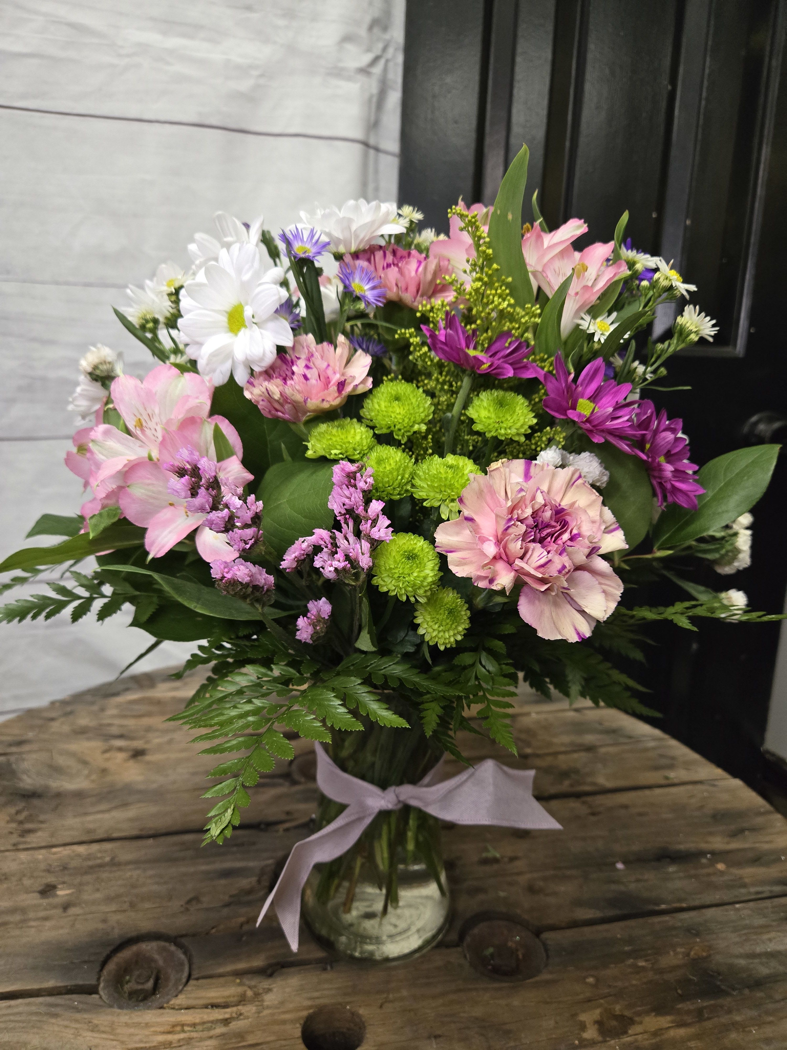 Purple Passion - .This is a lovely floral arrangement featuring a variety of colorful seasonal purple with a touch of pink flowers and greenery.  White daisies with yellow centers, adding a cheerful touch. Pink carnations with delicate ruffled petals, creating a soft and romantic feel. Purple chrysanthemums that bring depth and vibrancy to the arrangement. Bright green button mums, providing a fresh pop of color. Pink alstroemeria (Peruvian lilies) with subtle patterns, giving the bouquet a graceful look. Yellow solidago (goldenrod), offering a hint of texture and warmth. Complemented by lush green foliage, including ferns and other leafy accent. All flowers are subject to substations with seasonal availability.