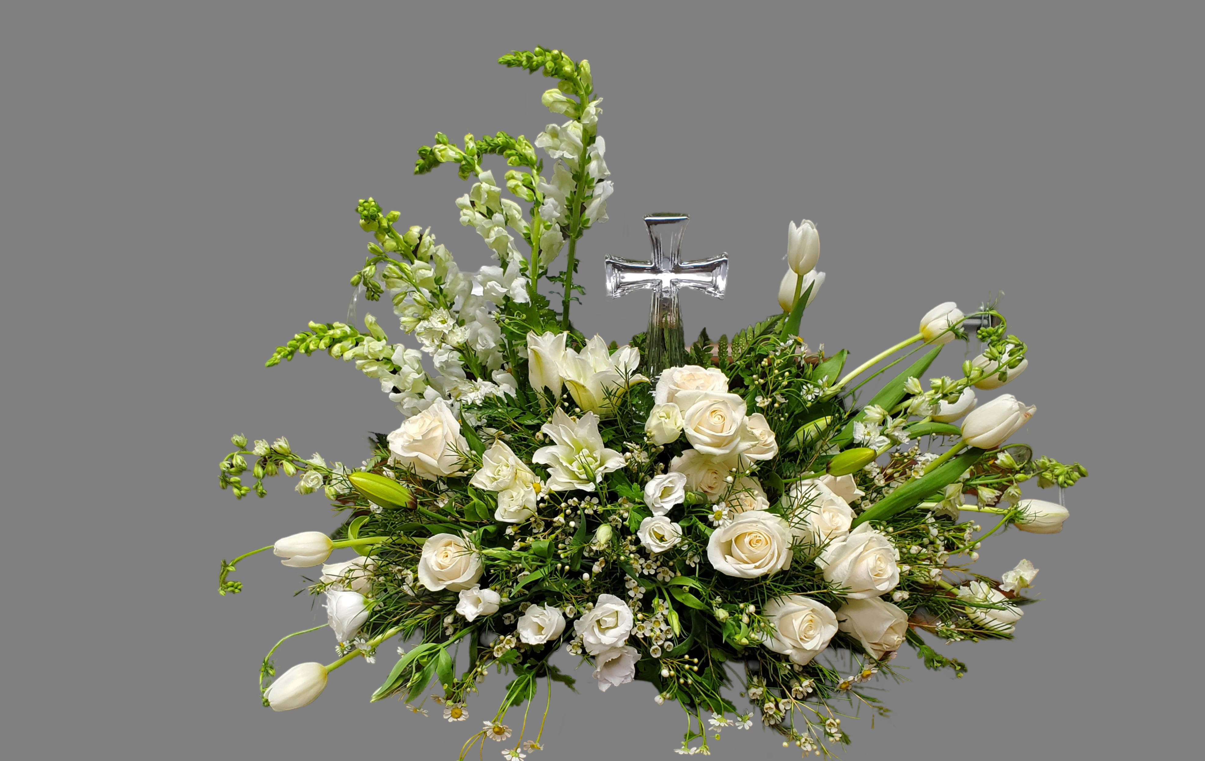 Serenity in White - This floral arrangement is a stunning and serene display of white blooms, symbolizing peace, purity, and grace. The centerpiece includes a glass cross, adding a religious or spiritual element, making it particularly suited for memorials or religious ceremonies. A balanced, symmetrical design with a focus on height provided by snapdragons and lilies. The arrangement cascades gently outward, creating a full, natural look. Overall, this piece is sophisticated, conveying a message of peace and hope, making it perfect for solemn and meaningful occasions. *All flowers are subject to substations with seasonal availability.