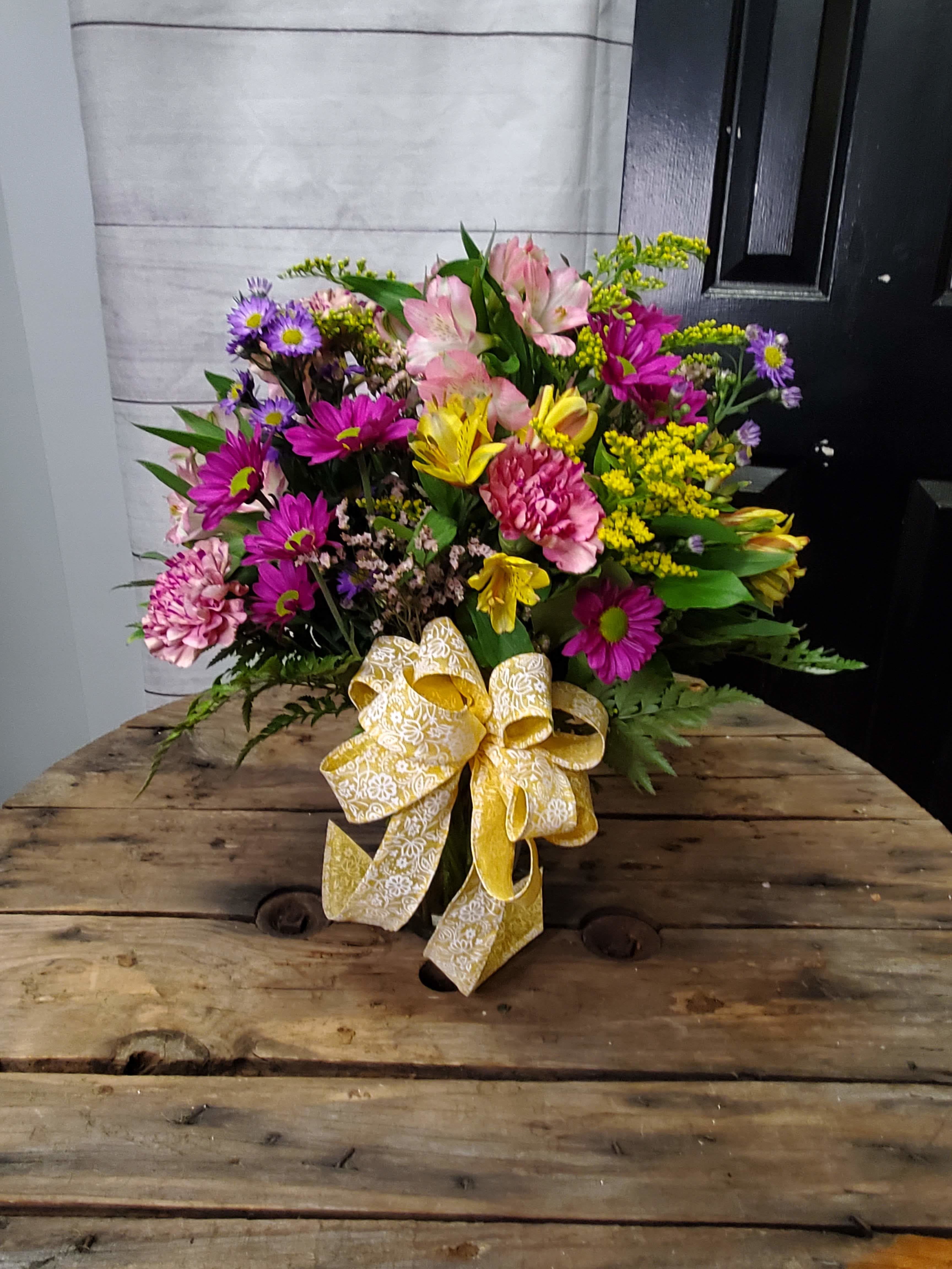 Simply Splendid  - This is a vibrant and cheerful floral arrangement that exudes energy and warmth.   Pink carnations and purple chrysanthemums add richness and depth to the bouquet with their bold, saturated colors. Yellow alstroemeria (Peruvian lilies) and solidago (goldenrod) brighten the arrangement with their sunny tones, adding a playful and lively contrast. Lavender daisies and small purple blooms provide softness and texture, complementing the bolder colors. Greenery, including lush ferns and leaves, creates a full, balanced backdrop and enhances the natural appeal. The arrangement is tied together with a large yellow ribbon, featuring a delicate white floral pattern, adding a charming and elegant finishing touch. This bouquet is perfect for celebrating joyful occasions or bringing a touch of happiness to any space. . All flowers are subject to substations with seasonal availability.