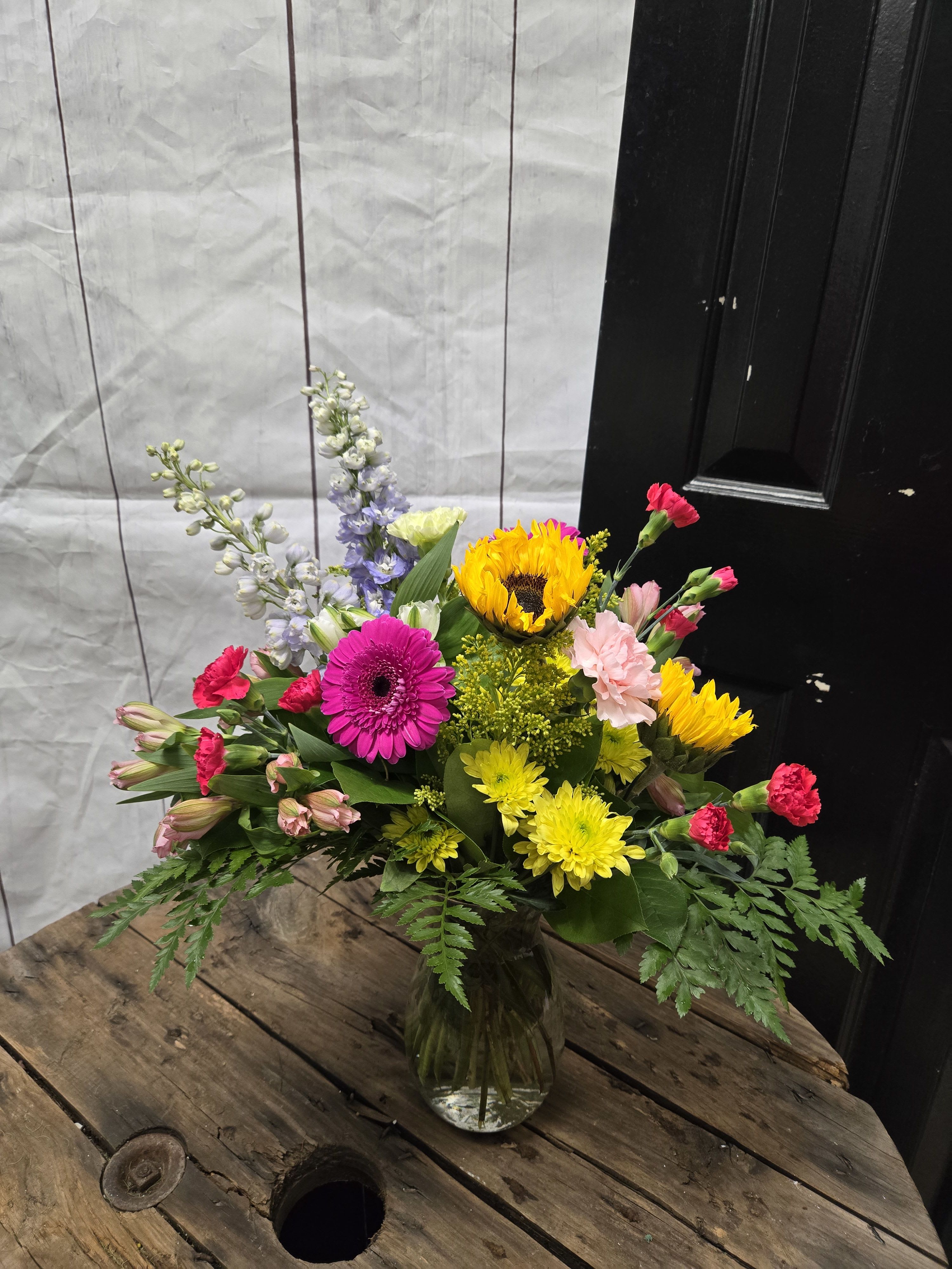 Sunlit Celebration - This arrangement is a lively and colorful bouquet that exudes joy and energy. It features:  Sunflowers, as the centerpiece, radiating warmth and happiness. Hot pink gerbera daisy, adding a bold and playful touch. Bright yellow mums, providing cheerful pops of color and texture. Soft pink carnations and alstroemeria, bringing elegance and a soft contrast to the bolder hues. Red mini carnations, adding vibrancy and depth. Delicate blue delphiniums, creating height and a subtle cool-toned accent. Lush greenery, such as ferns and leafy stems, to frame and complement the colorful blooms. The flowers are arranged in a clear glass vase. This bouquet has a fresh, celebratory feel, perfect for brightening any space or occasion! *All flowers are subject to substations with seasonal availability 