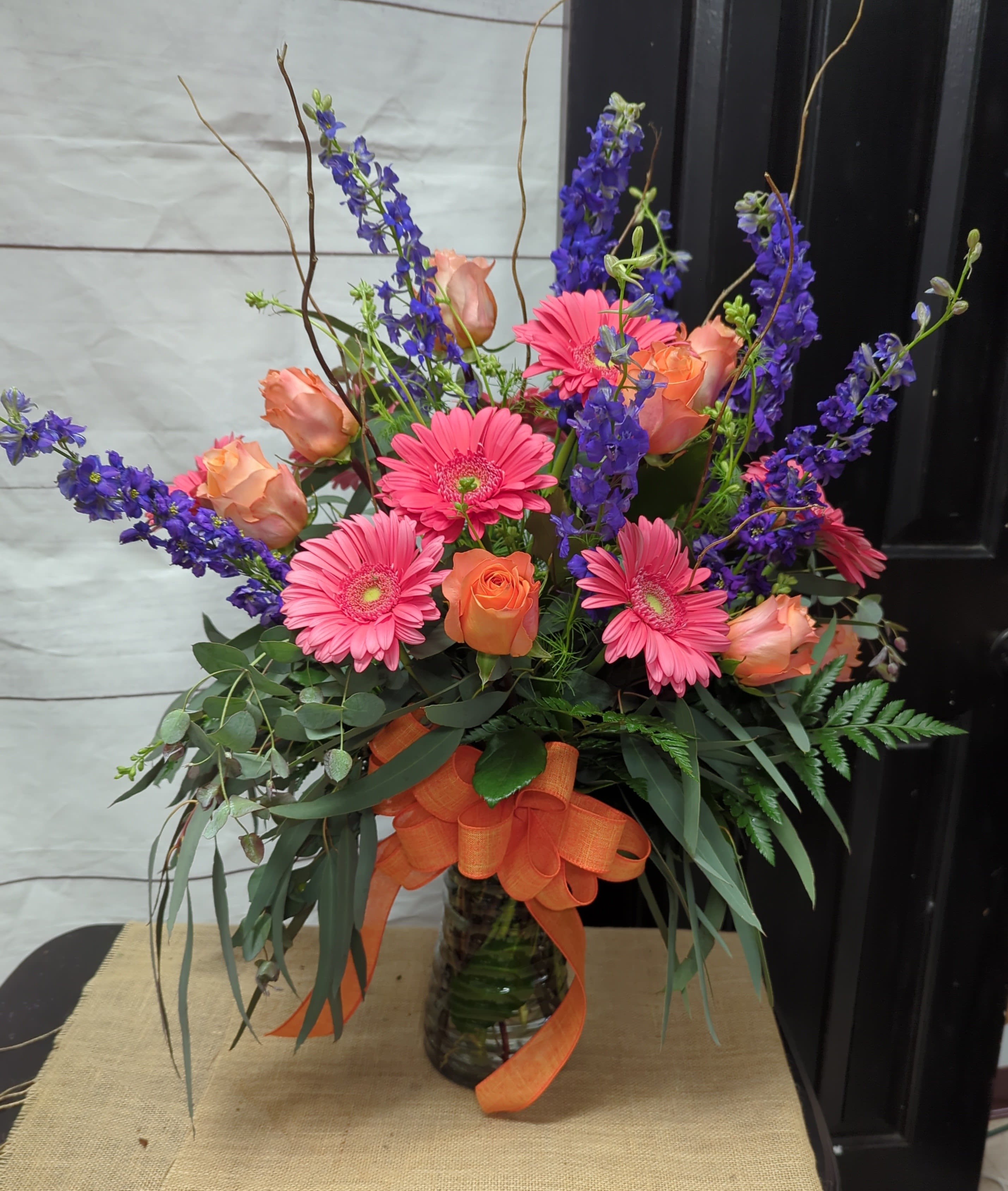 Vibrant Meadow Bliss - This arrangement bursts with energy and charm, featuring a lively combination of colors and textures. Bright pink gerbera daisies, creating a playful and cheerful focal point. Peach roses, offering warmth and softness with their delicate color. Deep blue delphiniums, adding vertical height and a touch of elegance. Rustic curly willow branches, providing a whimsical, natural aesthetic. Eucalyptus greenery and leatherleaf ferns, adding depth, texture, and a fresh, airy vibe. Tied with an orange bow and arranged in a clear vase, this design is perfect for celebrations, as a get-well gift, or to brighten any space with its joyful and vibrant presence