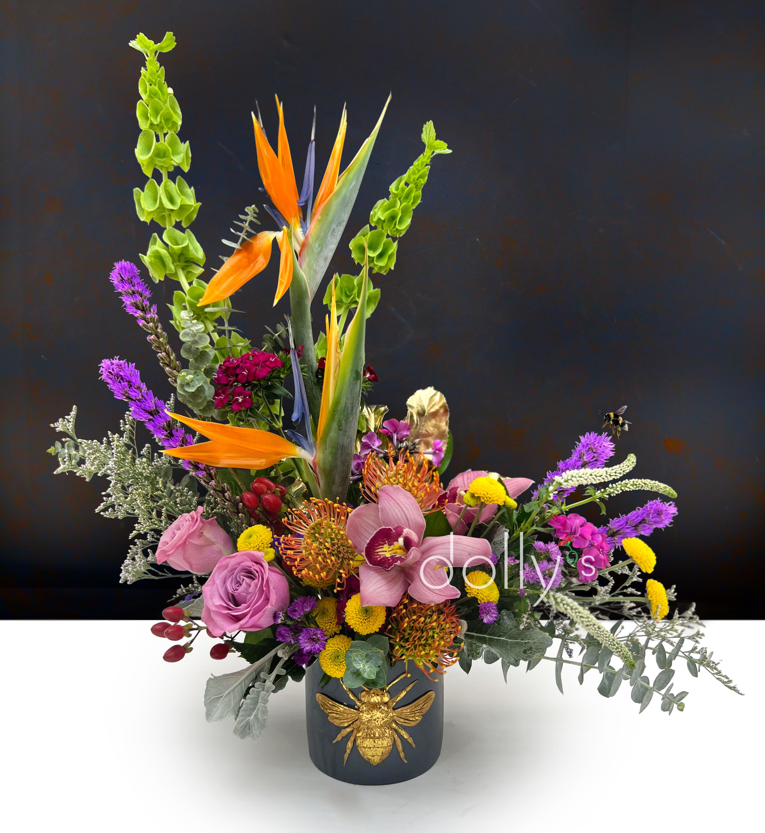 Queen Bee - This sovereign arrangement of cymbidium orchids,  pincushion protea, birds of paradise, liatris, and roses, each bloom chosen for its stately presence. Finished with gilded accents and presented in an opulent vase adorned with a grand golden bee, this composition reigns in elegance