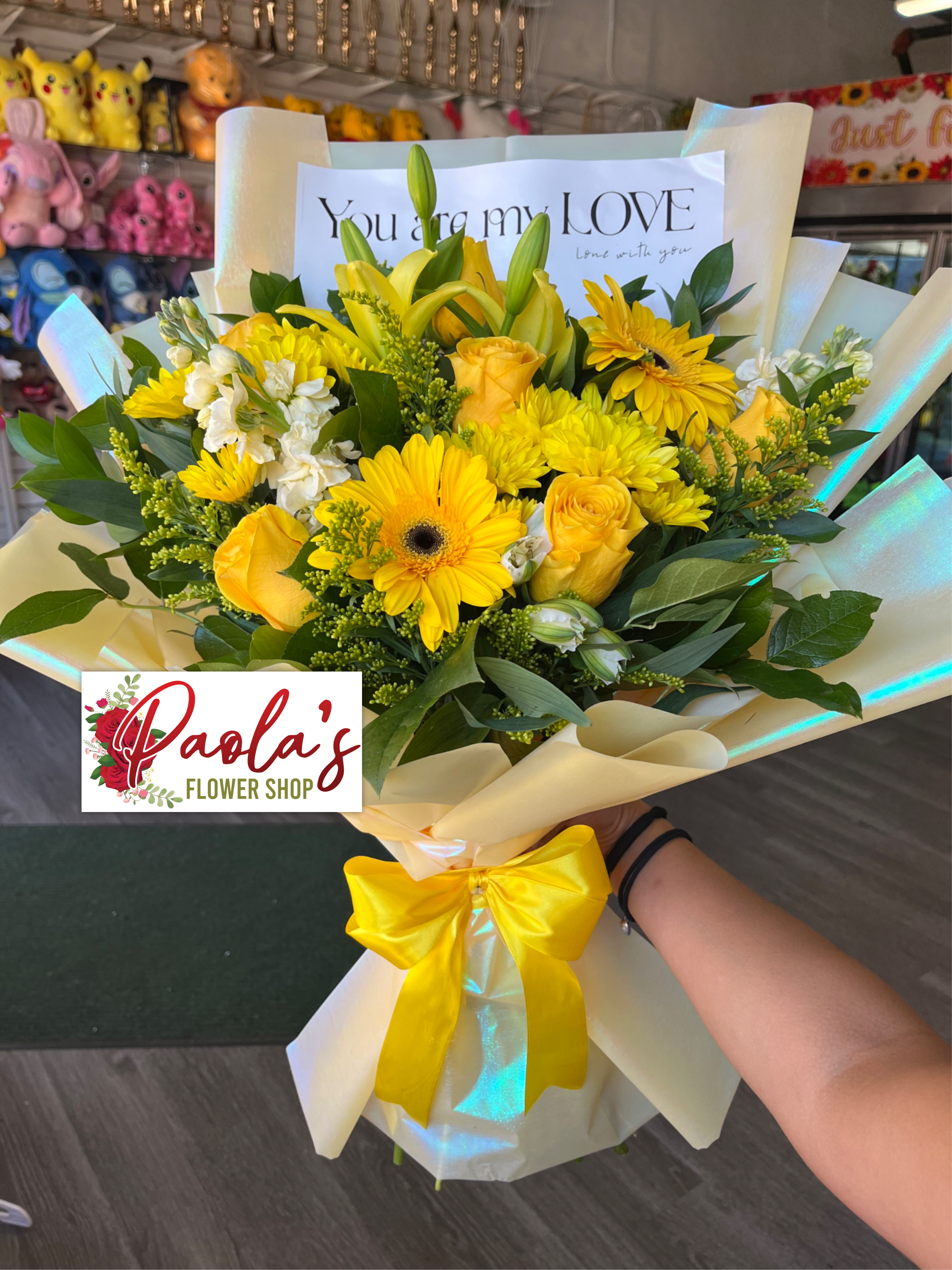 Golden Sunshine Mixed Bloom Bouquet - A cheerful hand-tied bouquet featuring a radiant mix of yellow blooms, including roses, gerbera daisies, chrysanthemums, and lilies, accented with fresh greenery. Wrapped in premium cream and iridescent paper and finished with a satin yellow ribbon, this arrangement is perfect for celebrations, congratulations, or simply brightening someone’s day.