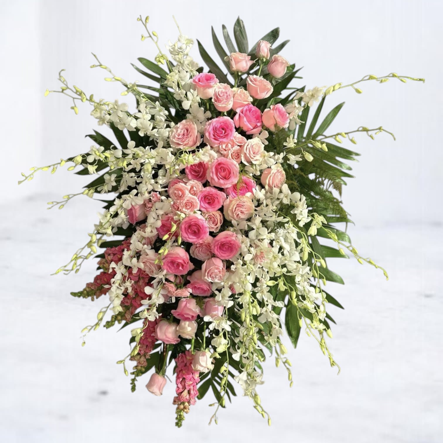 Rose Tribute Standing Spray - Honor a life of love and compassion with the Rose Tribute Standing Spray, a lush standing funeral spray designed to leave a lasting impression. Featuring layers of soft pink roses, cascading white dendrobium orchids, tropical greenery, and delicate snapdragons, this elegant design is perfect for memorial services, church ceremonies, or graveside tributes.  Thoughtfully arranged to evoke grace and serenity, this premium floral spray offers a beautiful way to express sympathy for the loss of a mother, daughter, sister, or cherished friend.  Substitution Disclaimer: We design each arrangement with intention. If any flowers become unavailable, we’ll substitute with blooms of equal value and visual impact while preserving the original color palette and essence.