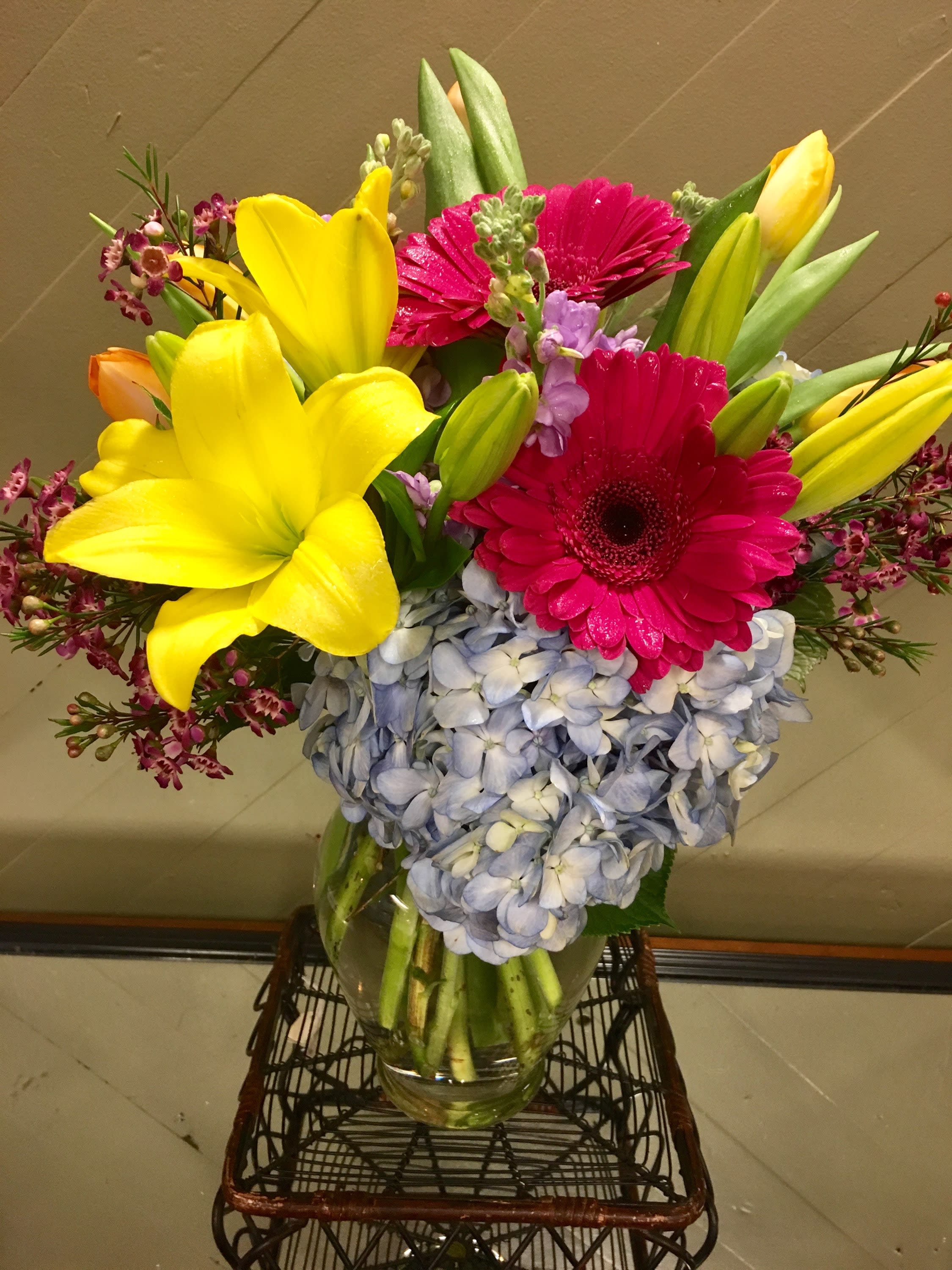 Happy Blooms - Cheery and bright bouquet with blue hydrangea, yellow lilies, hot pink gerberas and orange or pink tulips with waxflower. 