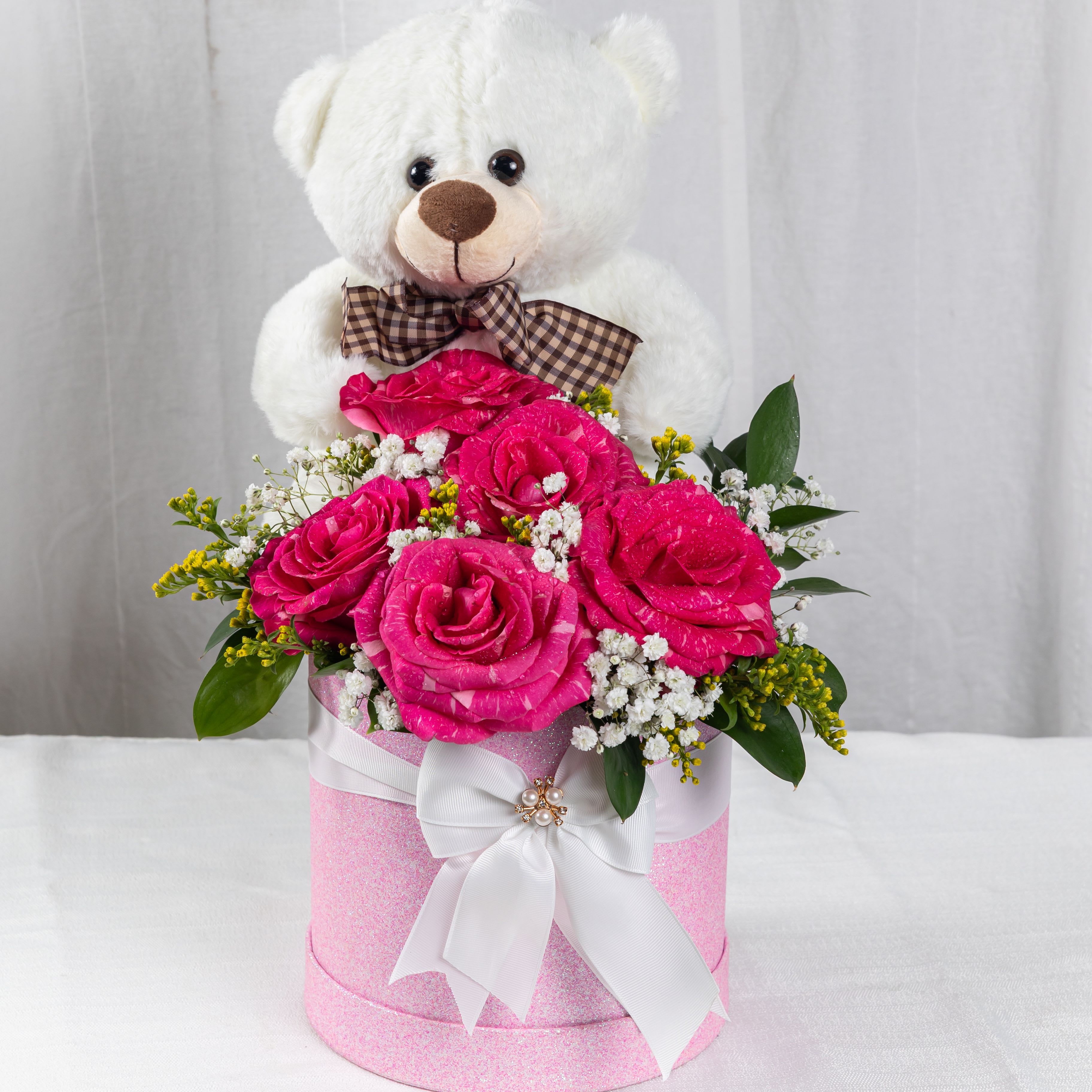 FLOWERS &amp; HUGS - A charming blend of vibrant pink roses, baby’s breath, and lush greenery nestled in a glittery pink container, crowned by a cuddly white teddy bear with a checkered bow. Finished with a pearl-accented ribbon, this arrangement is a tender gift of love and joy.
