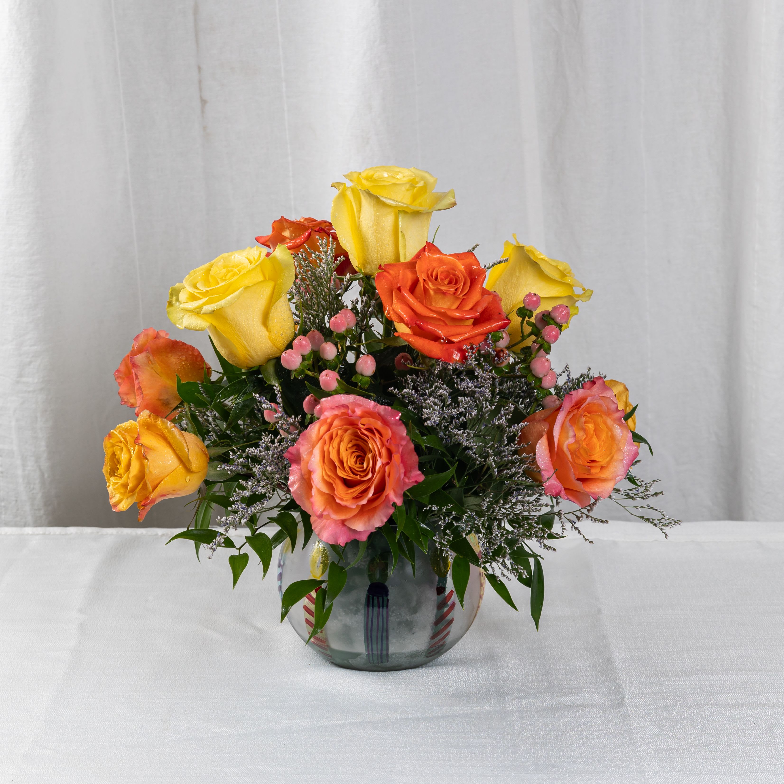 JOYFULL DAY - A radiant mix of yellow, orange, and pink roses with soft berries and white filler blooms, nestled in a clear glass vase. This cheerful bouquet brings warmth, elegance, and a touch of playful charm to any celebration.