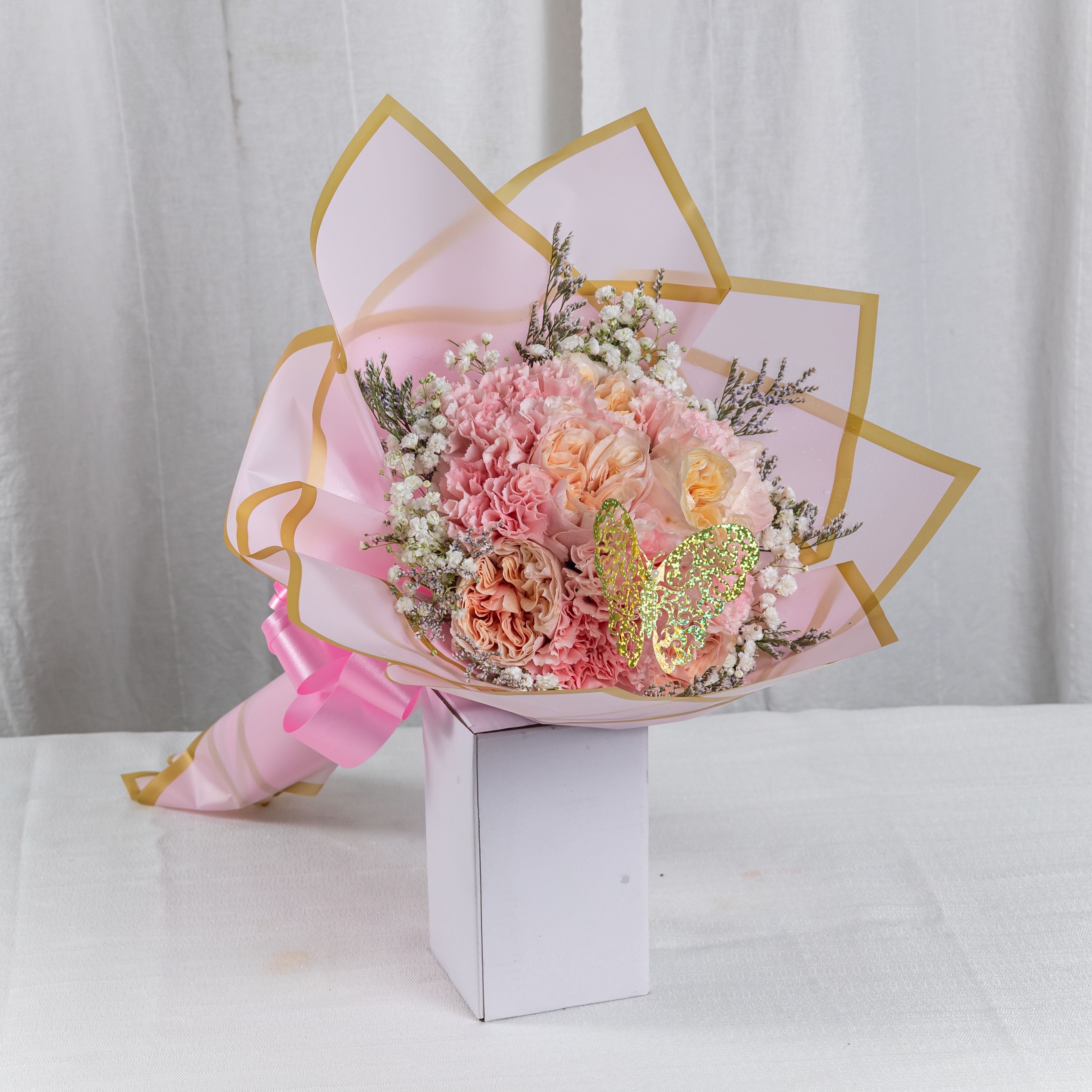 Petal Whispers - A tender bouquet of peach and pink carnations, blush roses, and lavender sprigs, wrapped in translucent pink paper with golden trim. Tied with a satin ribbon and adorned with a glittering butterfly, this arrangement evokes softness, celebration, and poetic charm.