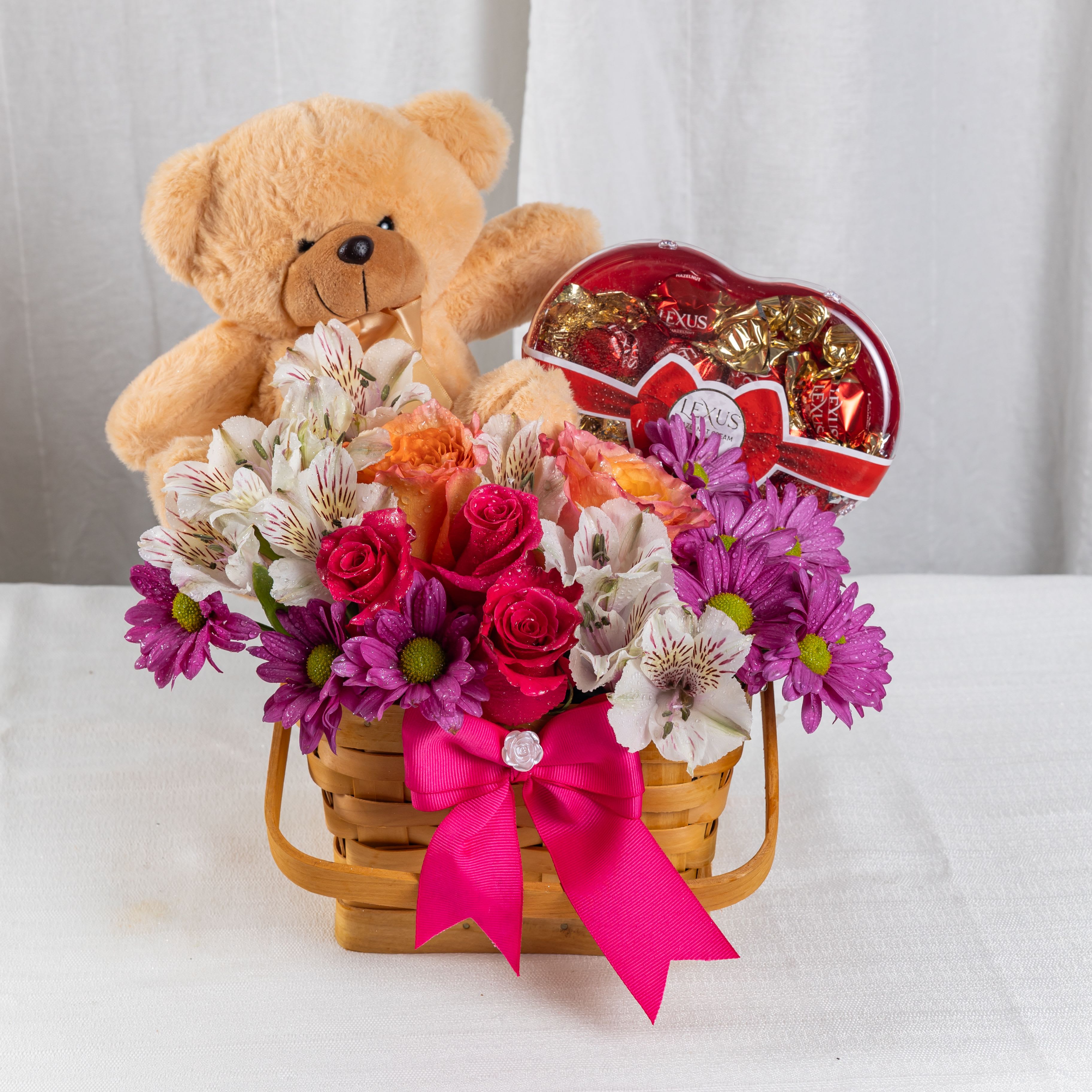 Smiles in Flowers - A heartwarming gift basket filled with pink daisies, alstroemerias, and orange roses, nestled beside a plush teddy bear and a heart-shaped box of Lexus chocolates. Tied with a jewel-accented ribbon, this charming ensemble celebrates affection, sweetness, and joyful surprises.