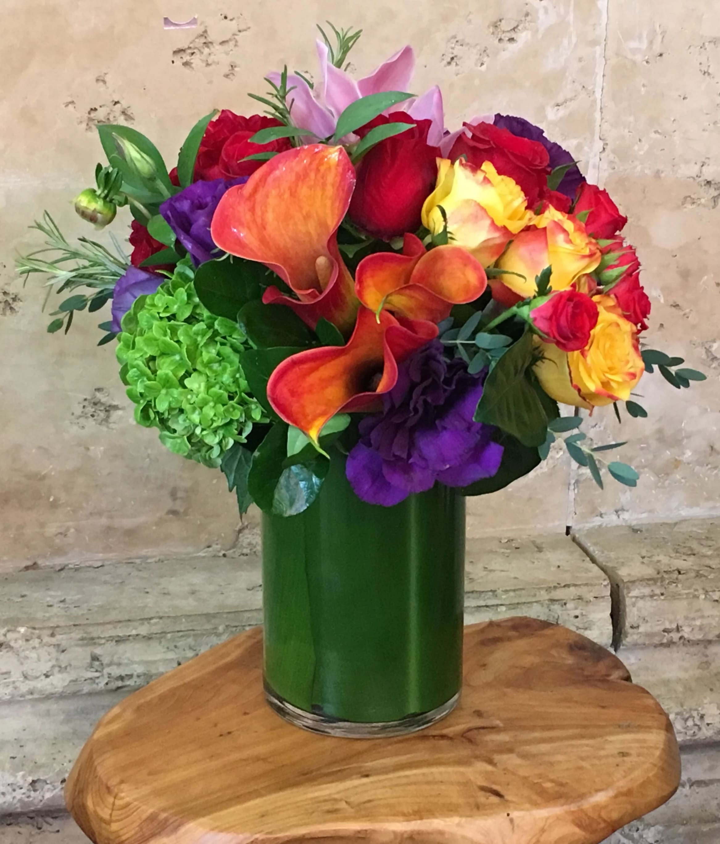 California - This arrangement feels like a celebration of pure color and movement.  A vibrant medley of fiery orange calla lilies, velvety red and sunset-yellow roses, rich purple lisianthus, and a fresh pop of green hydrangea—all gathered into a sleek emerald glass cylinder. The palette is bold yet harmonious, blending warmth and depth with unexpected contrast. Lush greenery softens the edges while the callas add sculptural drama, giving the piece a modern, artistic presence.  It’s playful, confident, and full of life—like a burst of sunshine with a touch of velvet evening. 