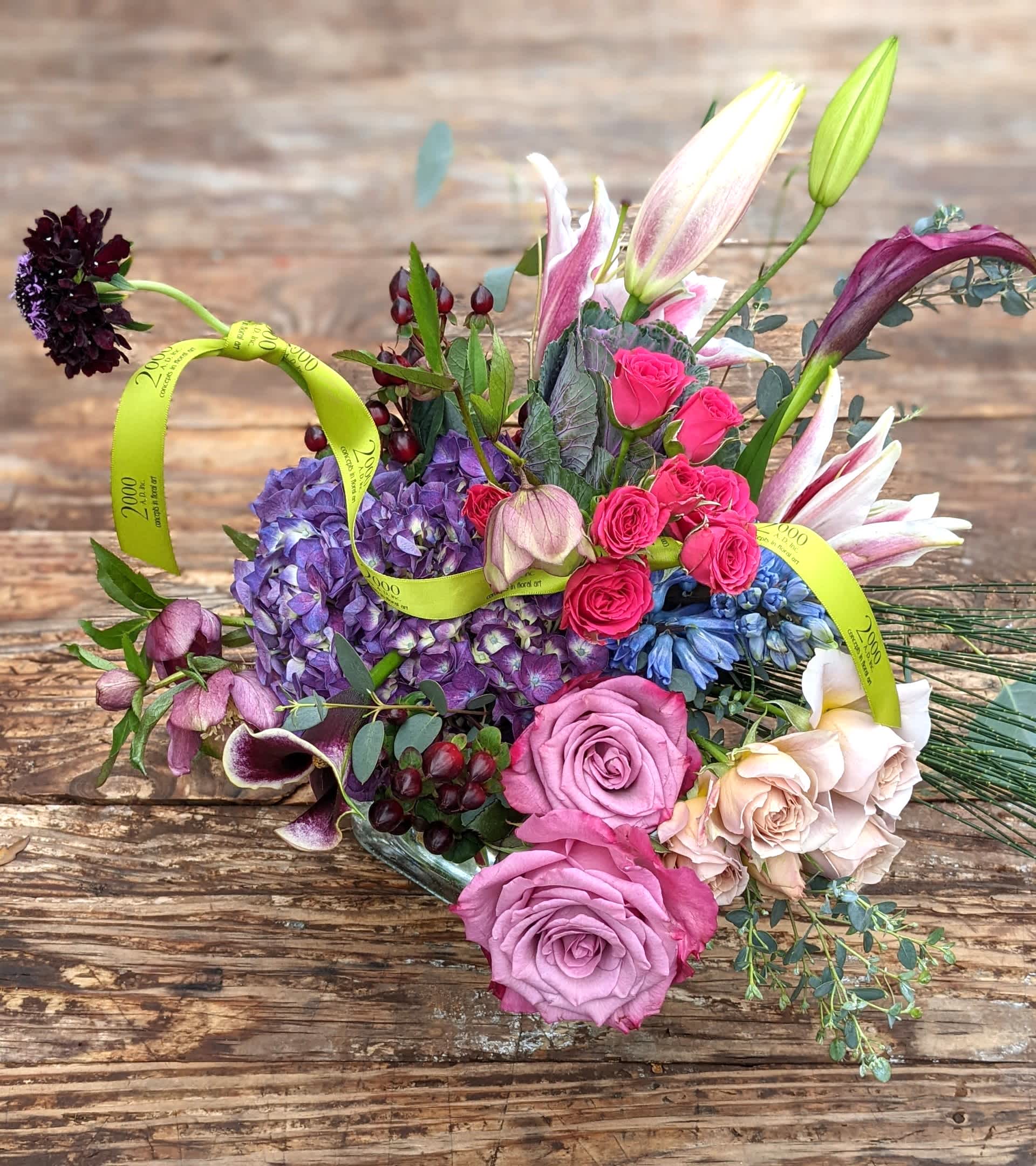 Love Is in the Air - Romantic palette of pinks purples and white with accent foliage. Presented in a clear glass container with exposed stems.
