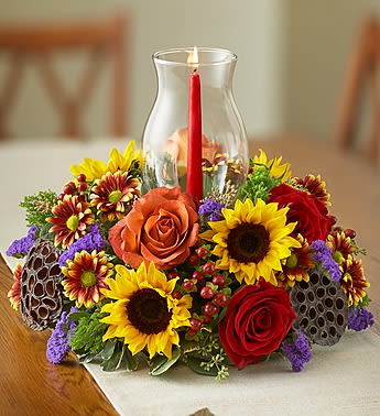 Harvest Glow Centerpiece - Product ID: 103584  EXCLUSIVE Cast a warm glow on every Fall gathering with this charming centerpiece arrangement. A hand-designed medley of bi-color roses, sunflowers, mini green hydrangeas and more are gathered together around a glass hurricane with a tapered candle to set the table in truly original style. Fresh arrangement of bi-colored roses, red hypericum, purple statice, white kale, sunflowers, green mini hydrangeas and variegated pittosporum Centered around a clear glass hurricane; includes a 12âH taper candle Clear glass hurricane measures 8.75âH Large arrangement measures approximately 13"D Medium arrangement measures approximately 12"D Small arrangement measures approximately 11"D Our florists select the freshest flowers available, so colors, varieties and container may vary due to local availability