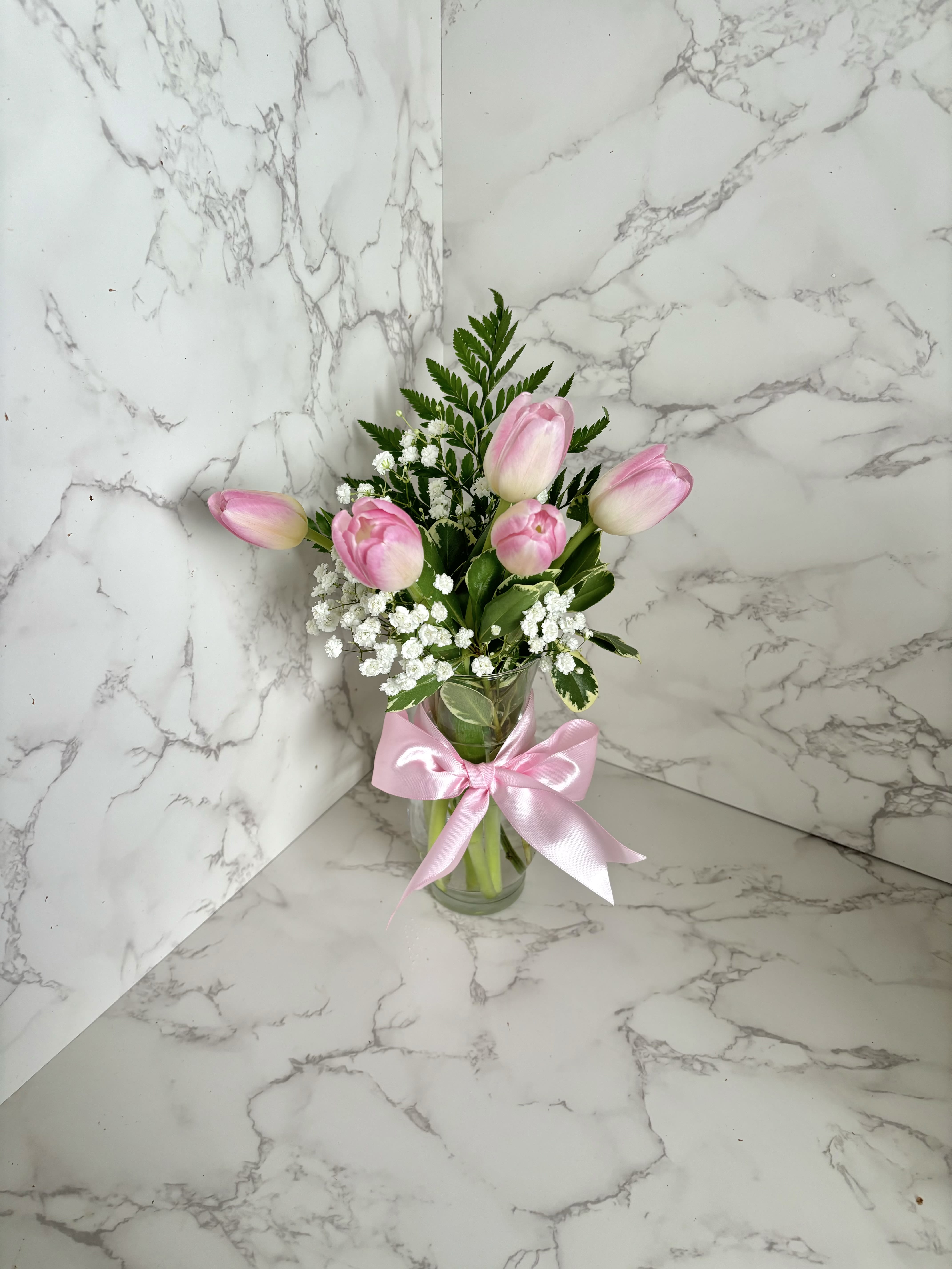 Blush Elegance Tulips in Glass Vase - Simple, sweet, and full of charm, this petite tulip arrangement brings a touch of elegance to any space. Featuring fresh tulips accented with delicate baby’s breath and lush greenery, it’s presented in a clear glass vase tied with a satin ribbon bow. Perfect as a thoughtful gesture, desk accent, or small celebration gift.  Arrangement includes: 	•	5 fresh tulips (colors may vary based on availability – pink, purple, white, or yellow) 	•	Baby’s breath (Gypsophila) 	•	Assorted greenery 	•	Clear glass vase with coordinating satin ribbon  A lovely choice for birthdays, thank-you gifts, or just because—it’s beauty in its most graceful form.
