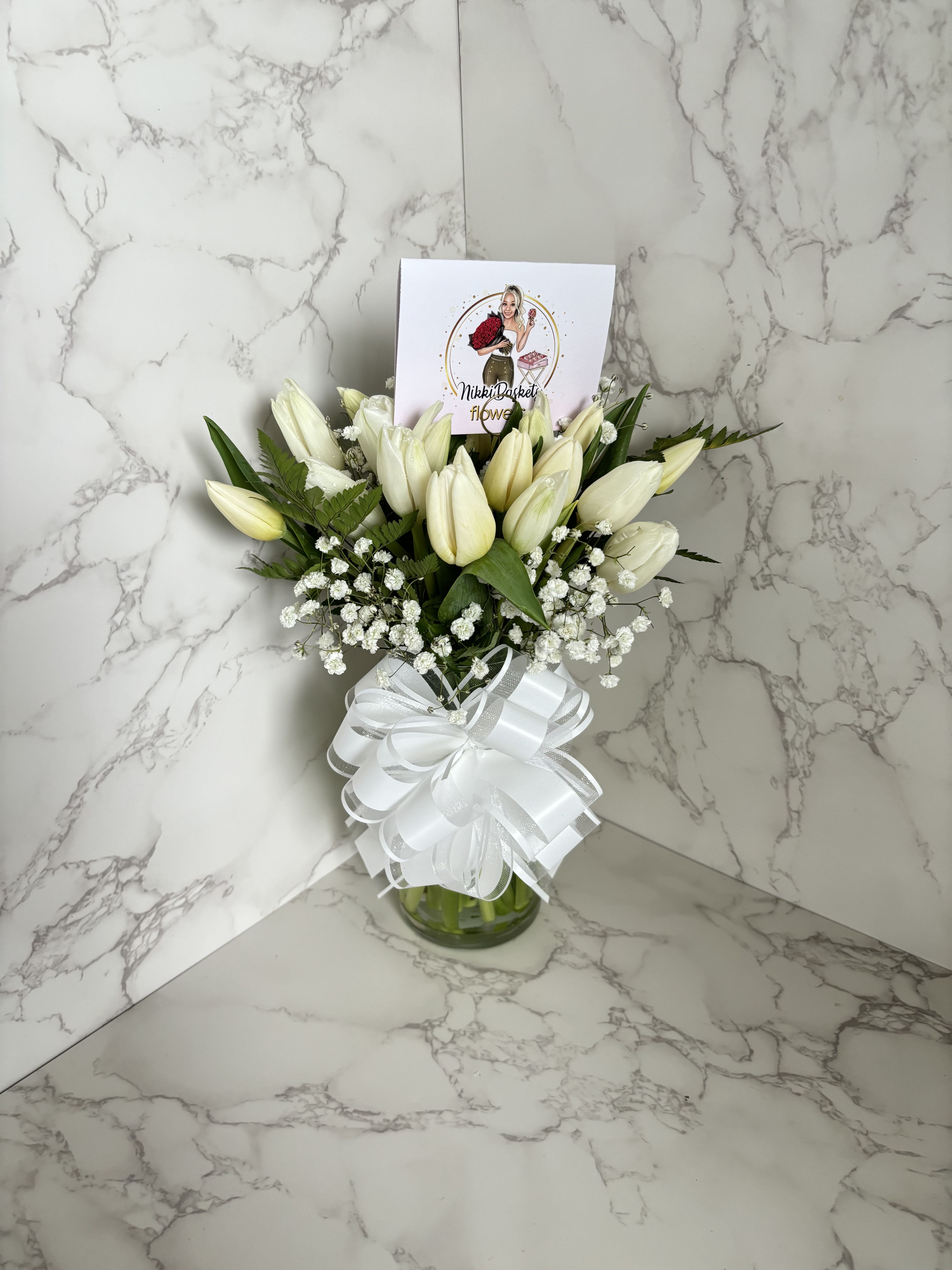 Elegant White Tulips In Vase (20) - A timeless arrangement of fresh white tulips, delicately accented with baby’s breath and lush greenery, beautifully displayed in a clear glass vase. Finished with a large white bow for an elegant touch, this piece radiates purity, grace, and sophistication. Perfect for weddings, anniversaries, sympathy gestures, or simply brightening someone’s day with classic beauty.