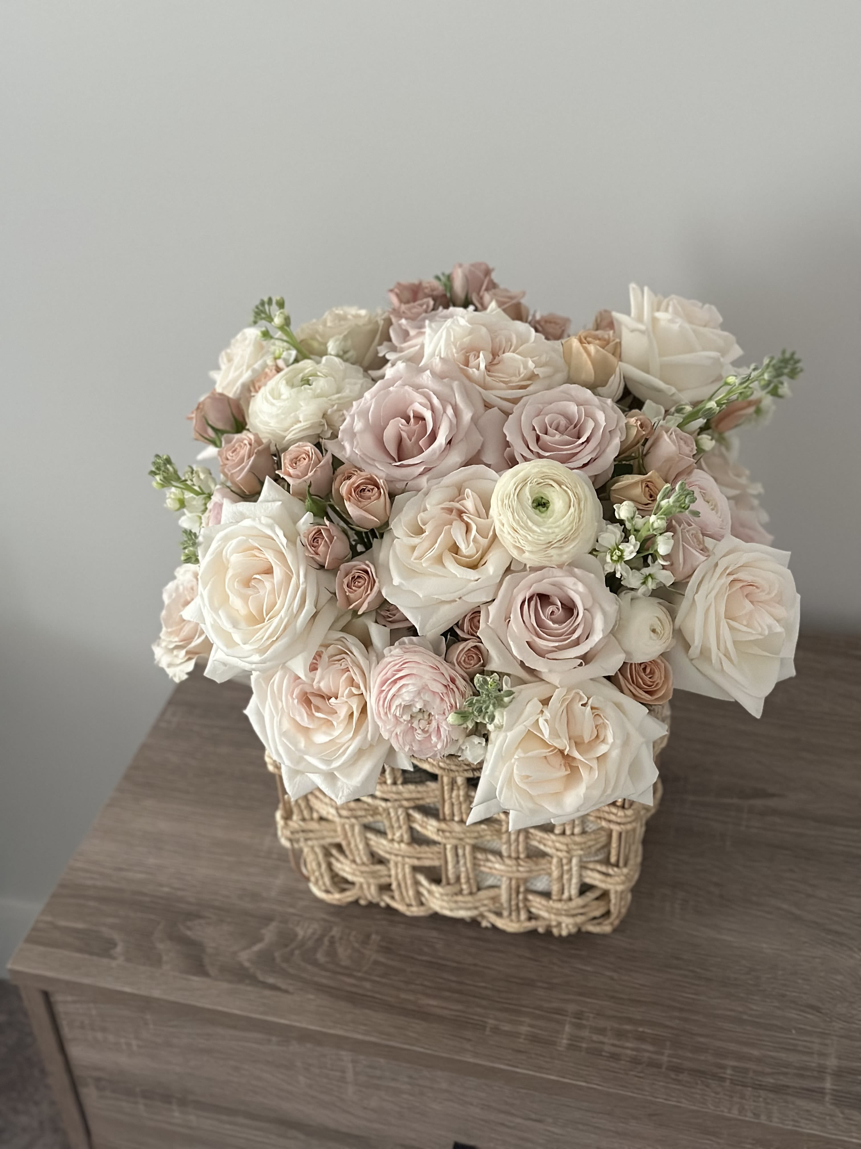 Blush Basket - A charming arrangement of soft blush and ivory roses, delicate ranunculus, and tender buds, nestled in a rustic woven basket. This bouquet is a gentle whisper of beauty and grace — ideal for cozy celebrations, home décor, or gifting love in its purest form.
